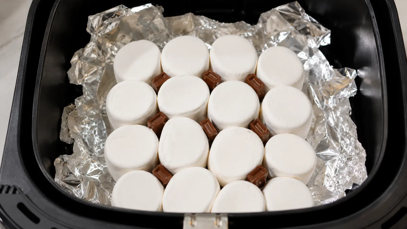 Overhead view of marshmallows and chocolate pieces on foil inside an air fryer as a hand places another chocolate piece.