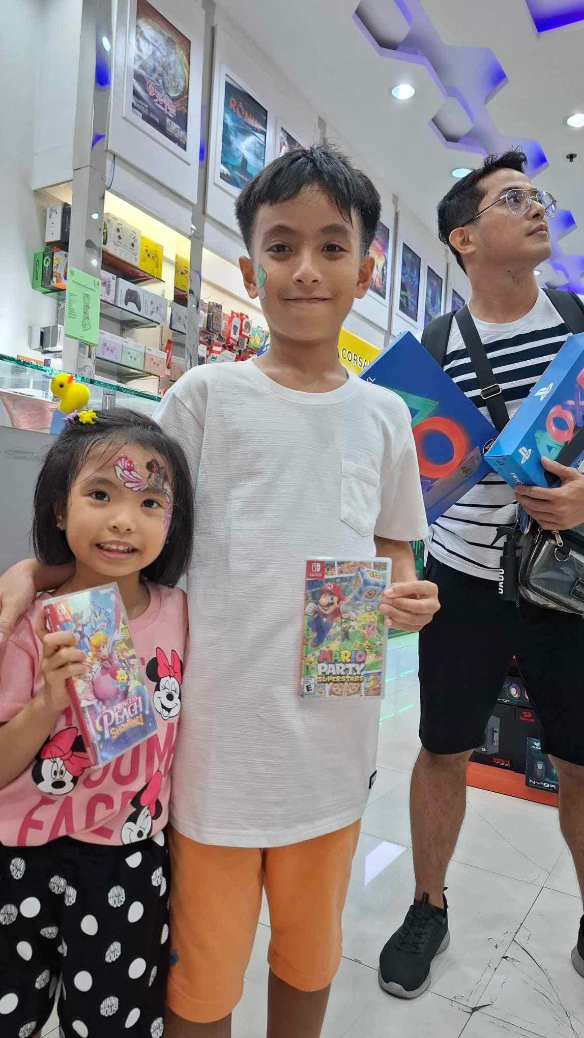 A photo of my two kids. My daughter on the left is holding a princess peach game. My son in the middle is holding a Mario Party Superstars game, and me on the right most holding a Playstation LED accessory