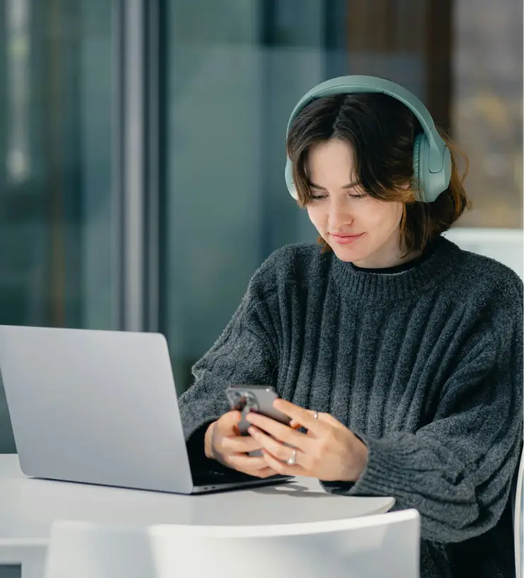 A person wearing headphones sits at a table, looking at their phone, with a laptop open in front of them. Natural light filters through a window.