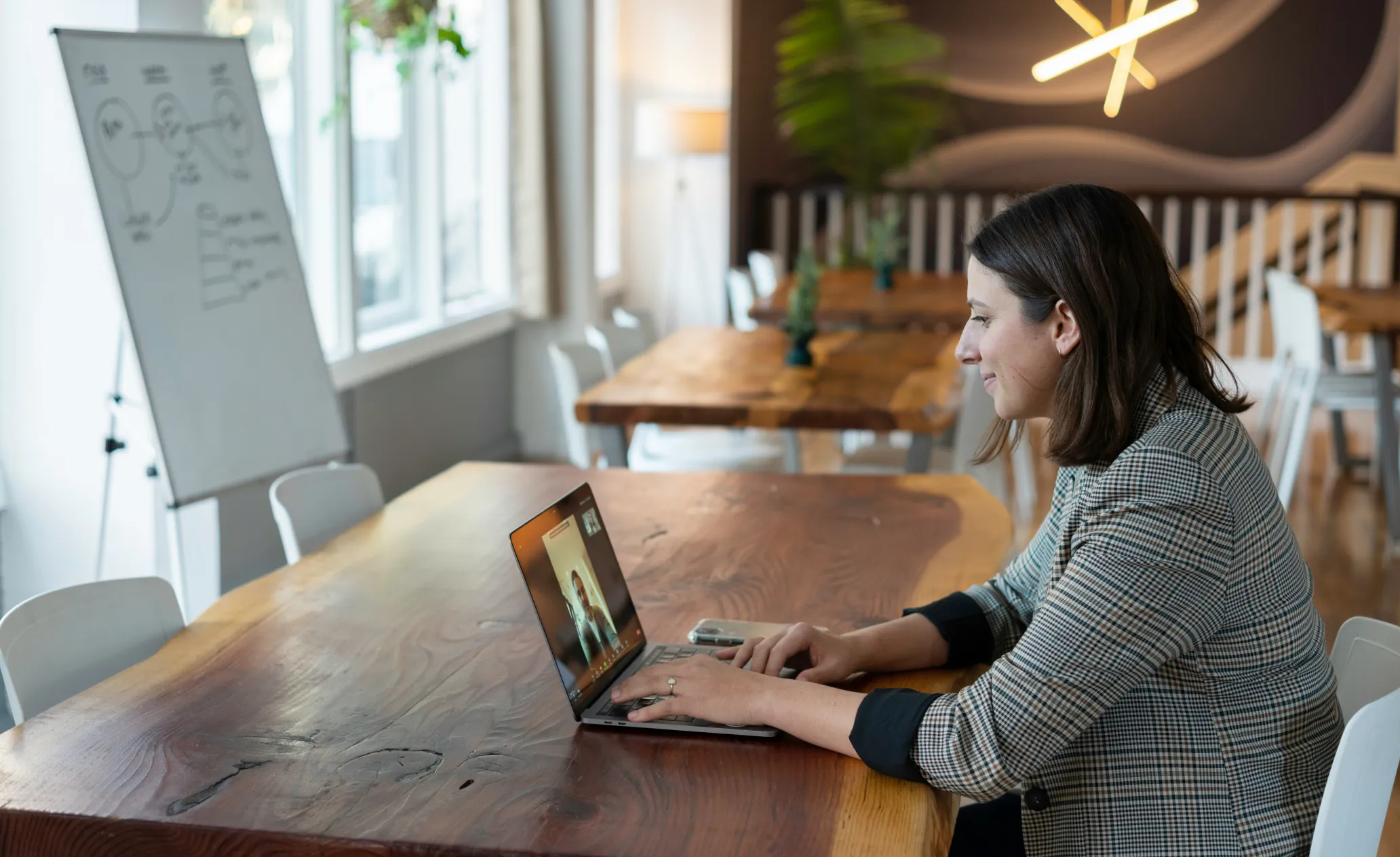 A woman in a checkered blazer sits at a wooden table, engaging in a video call on her laptop in a bright, modern workspace.