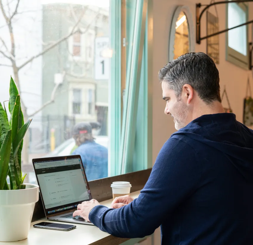 A person works on a laptop in a coffee shop, with a potted plant nearby and a takeaway coffee cup on the counter.