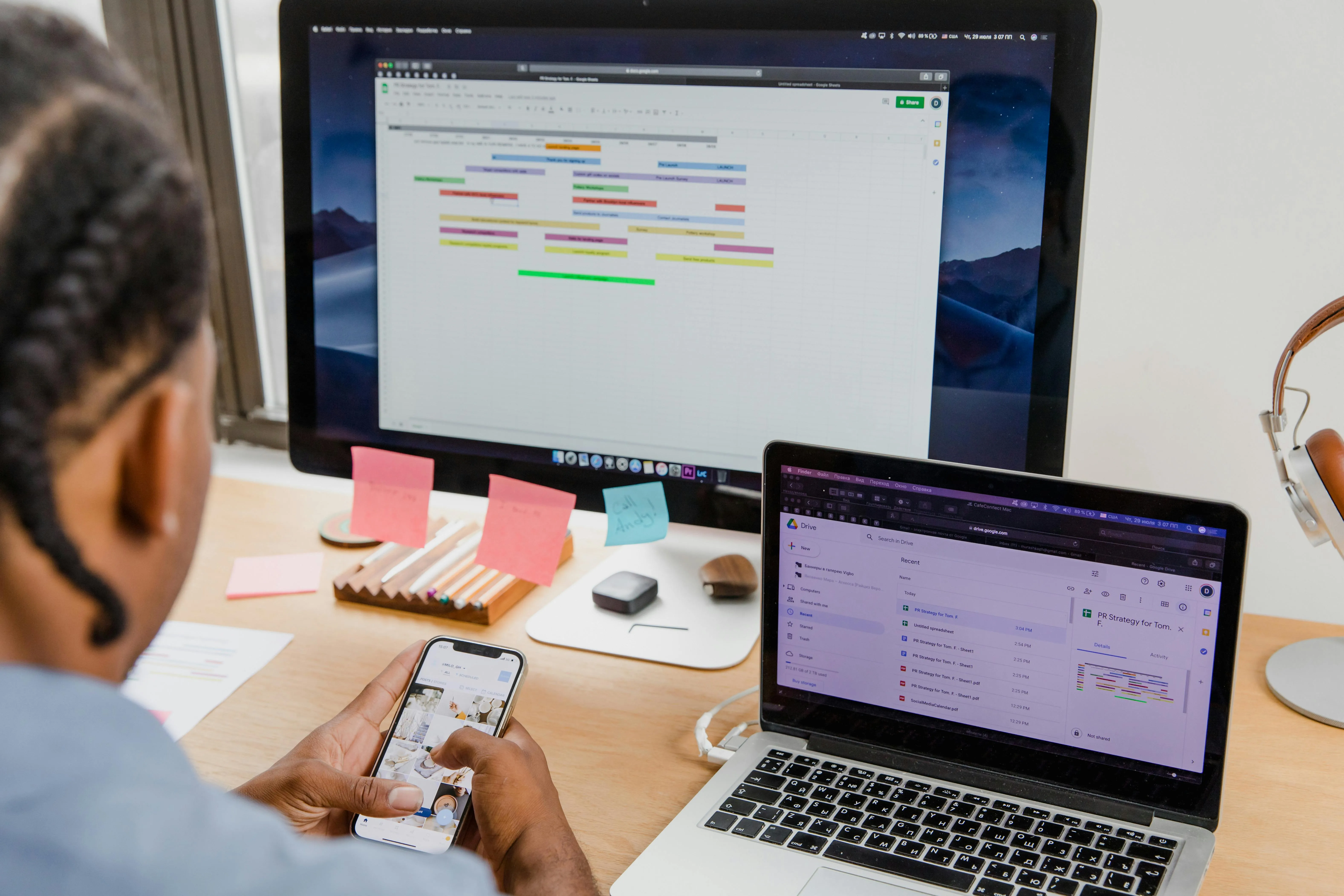 Remote worker sitting at a desk using a smartphone, laptop, and large monitor with project management and cloud storage tools open.