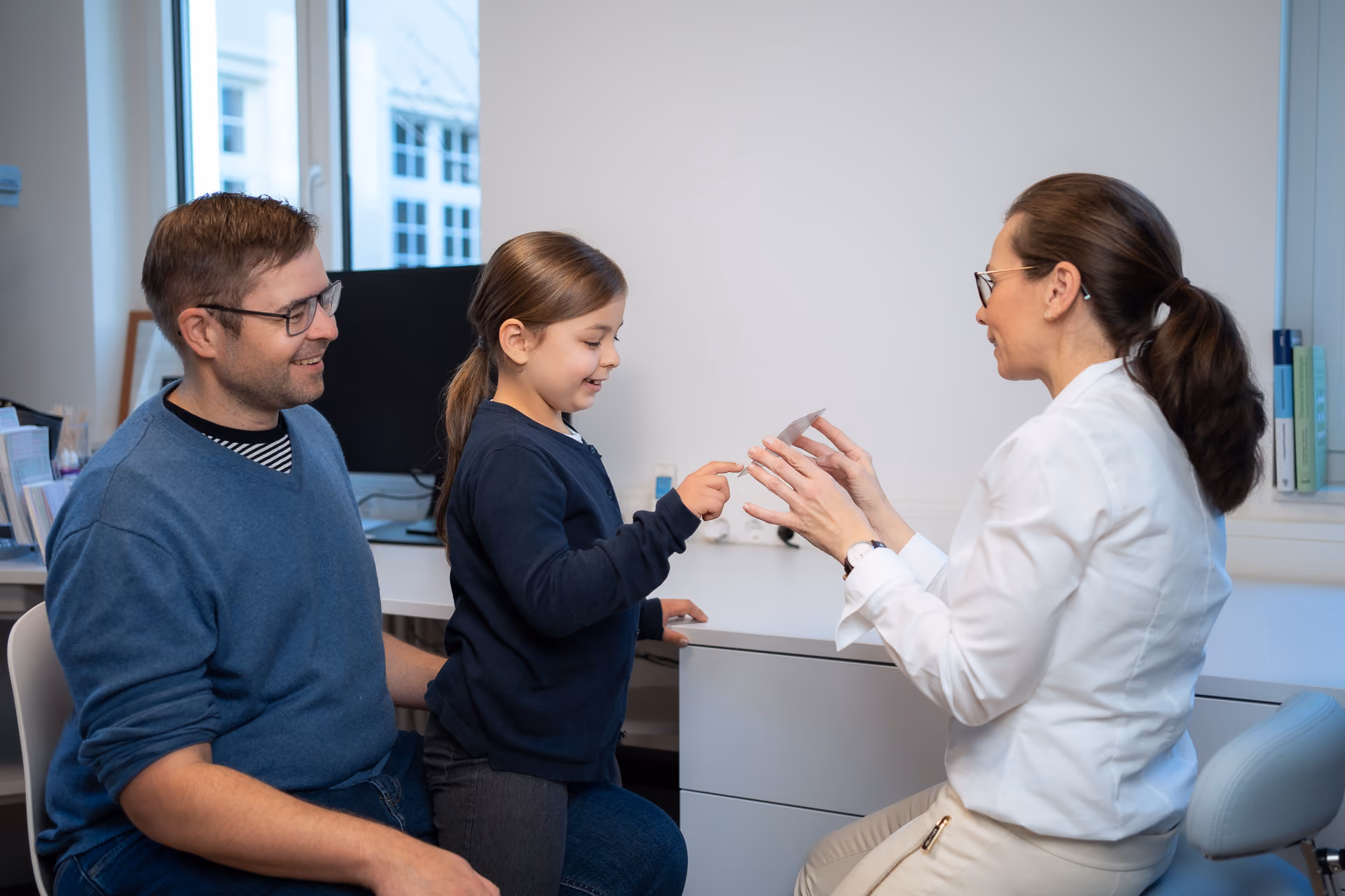Ärztin bespricht Untersuchungsergebnisse und Therapiemöglichkeiten mit 
  Eltern und Kind im Sprechzimmer.