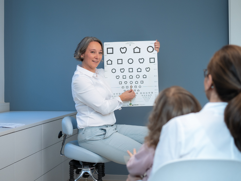 Orthoptistin zeigt Kindern eine Tafel mit verschiedenen Symbolen und 
  Formen zur Überprüfung der visuellen Wahrnehmung.