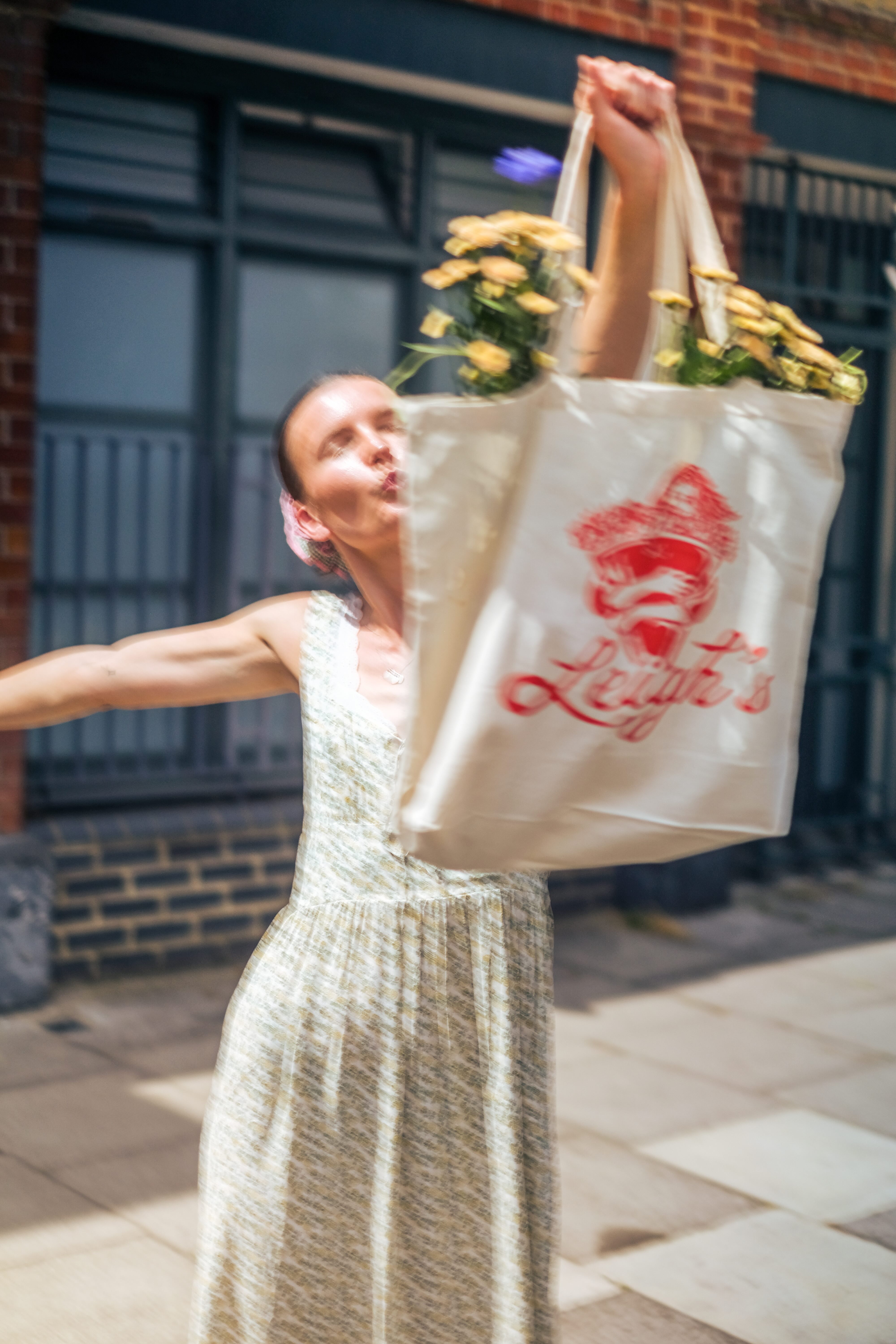 Woman in a light floral dress holding up a tote bag filled with yellow flowers on a sunny street.