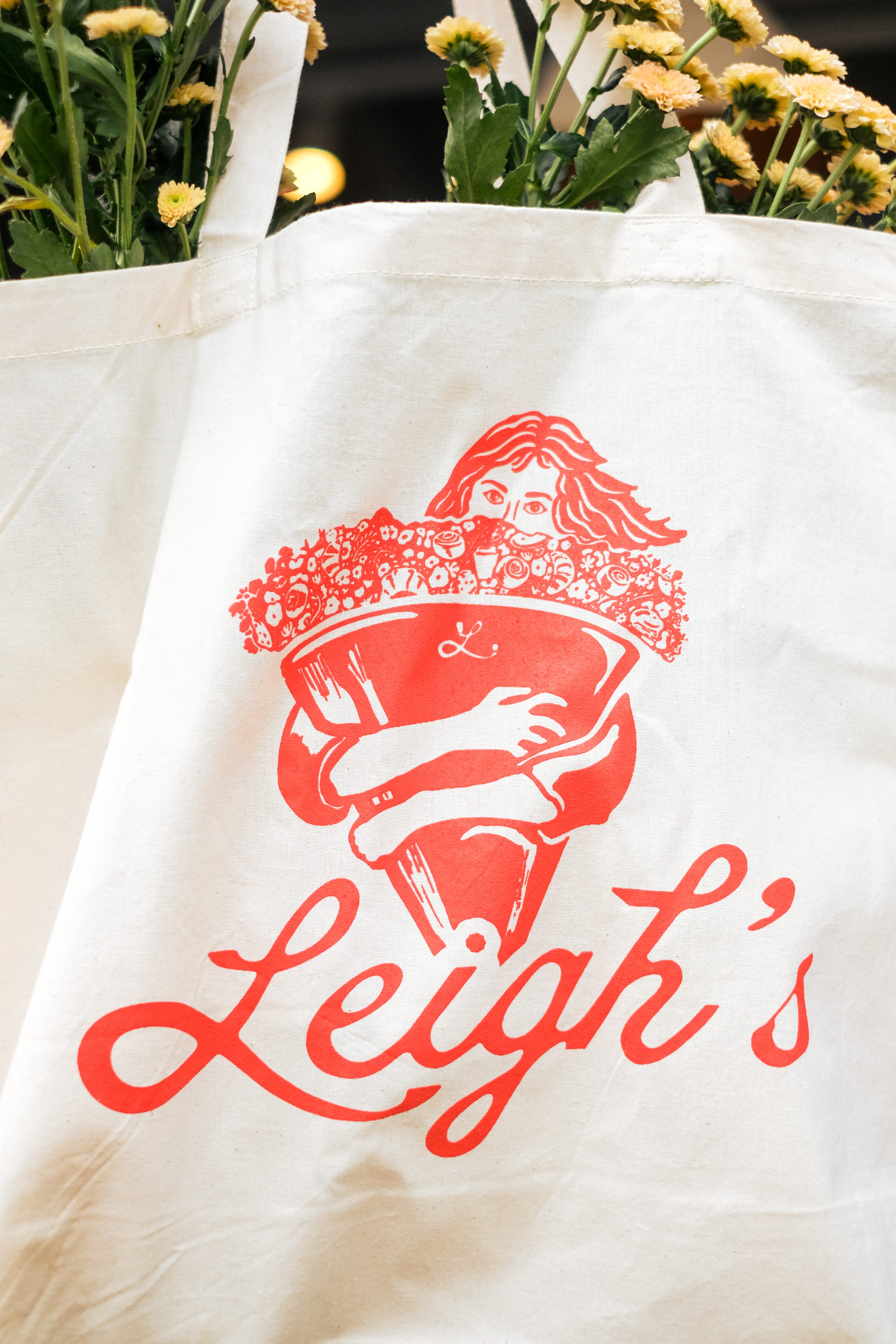 Canvas tote bag with red illustration of a person hugging a large bouquet of flowers and the text 'Leigh's'.