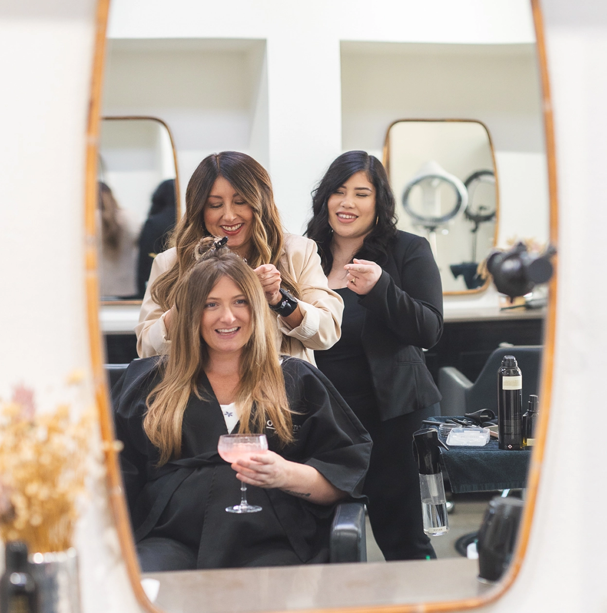 A woman with long blonde hair sits in a salon chair smiling and holding a drink while a stylist and an assistant work on her hair reflected in a mirror.