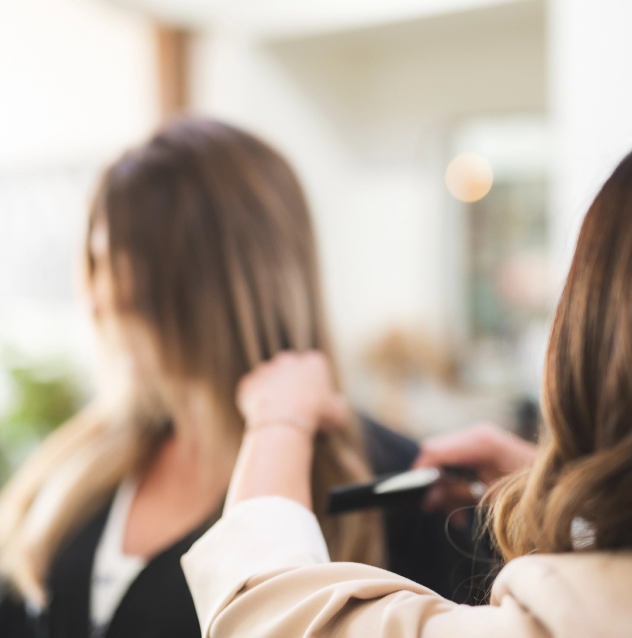 Hairdresser combing long hair of a seated woman in a salon with blurred background.