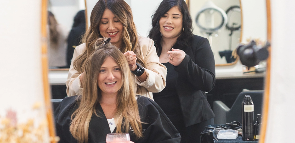 Hair stylist smiling while working on a seated woman's hair in a salon, another woman watches and smiles.