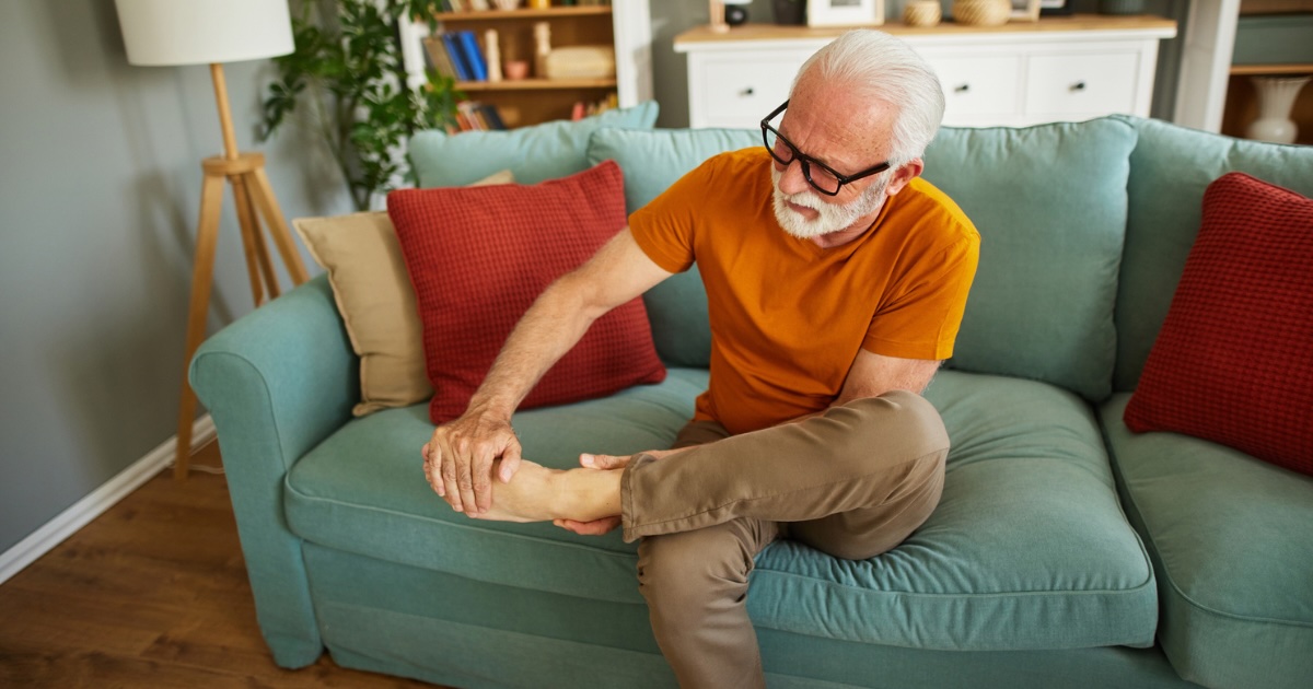 An elderly man with diabetes sitting on a couch holding his foot in pain, suffering from diabetic neuropathy effects.