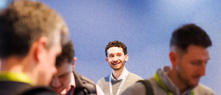 Smiling man with curly hair and lanyard standing against a blue wall, people blurred in foreground.
