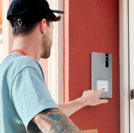 Man in a black cap and blue shirt interacting with a wall-mounted smart doorbell on a red wall near a door.