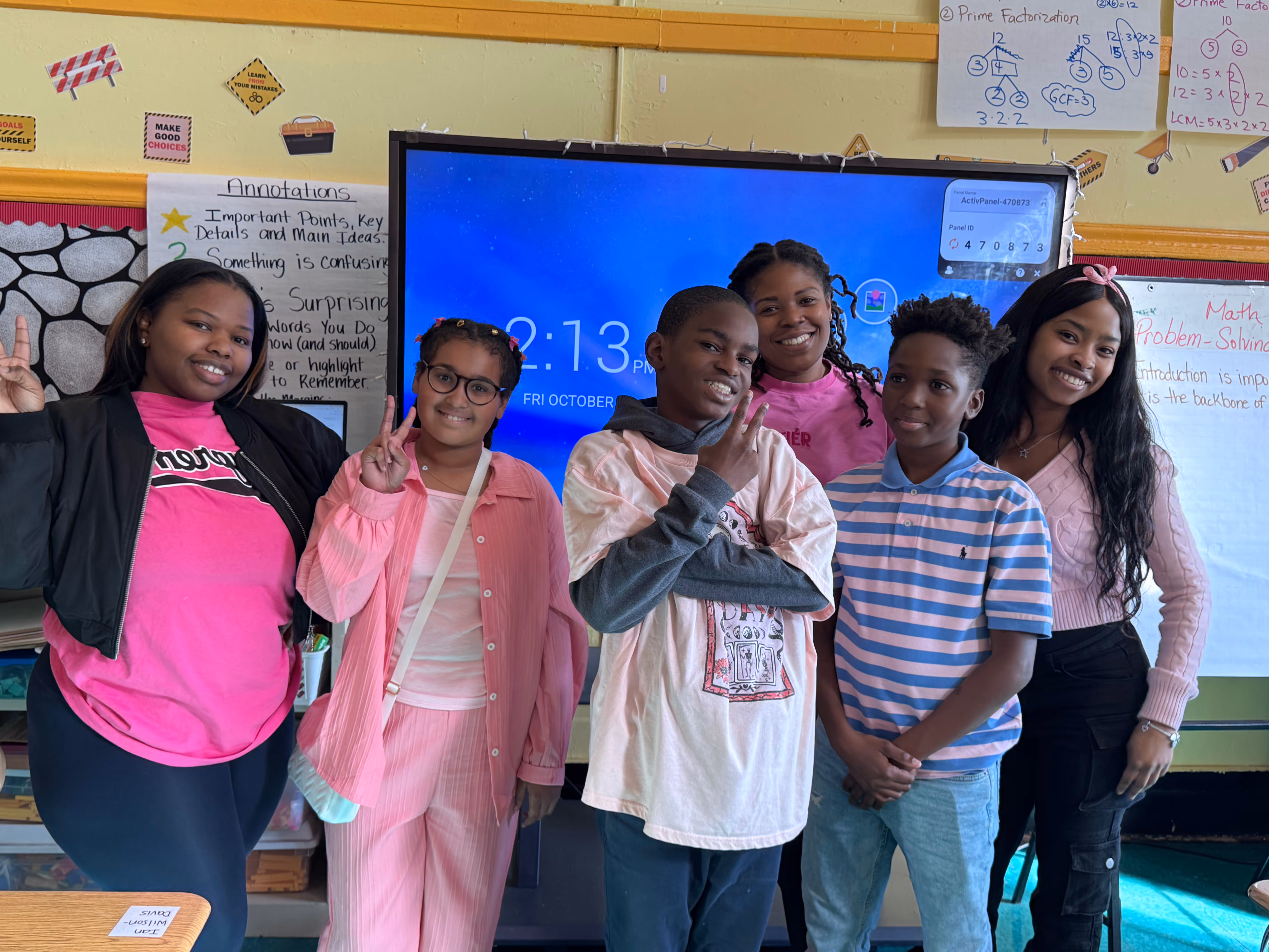 "Six students standing in front of a smart board in a classroom, smiling and making peace signs. Many are wearing pink for what appears to be a themed day. The background includes educational posters and math charts."