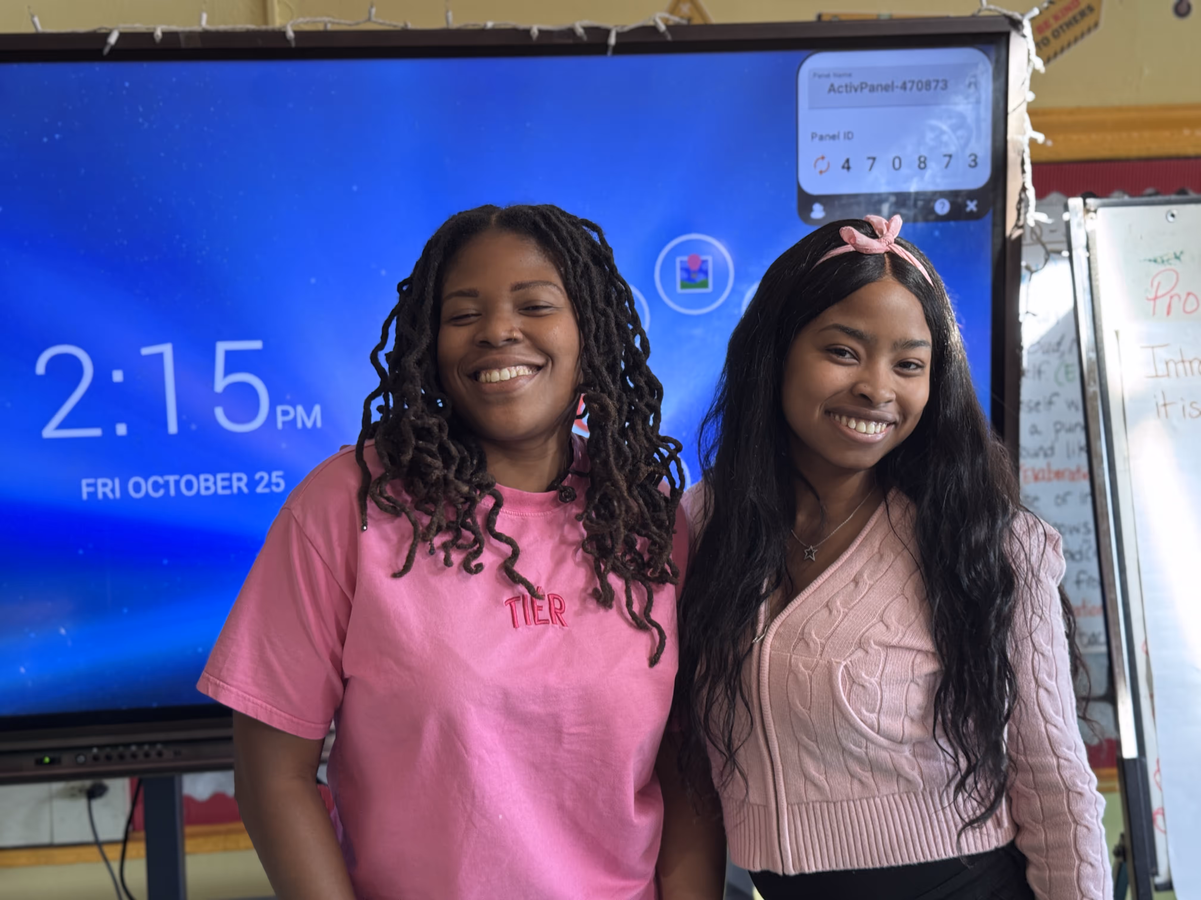 Two women smiling and standing in front of a large digital classroom display showing the time "2:15 PM" and date "Friday, October 25." One is wearing a pink T-shirt, the other a pink sweater with a bow headband.