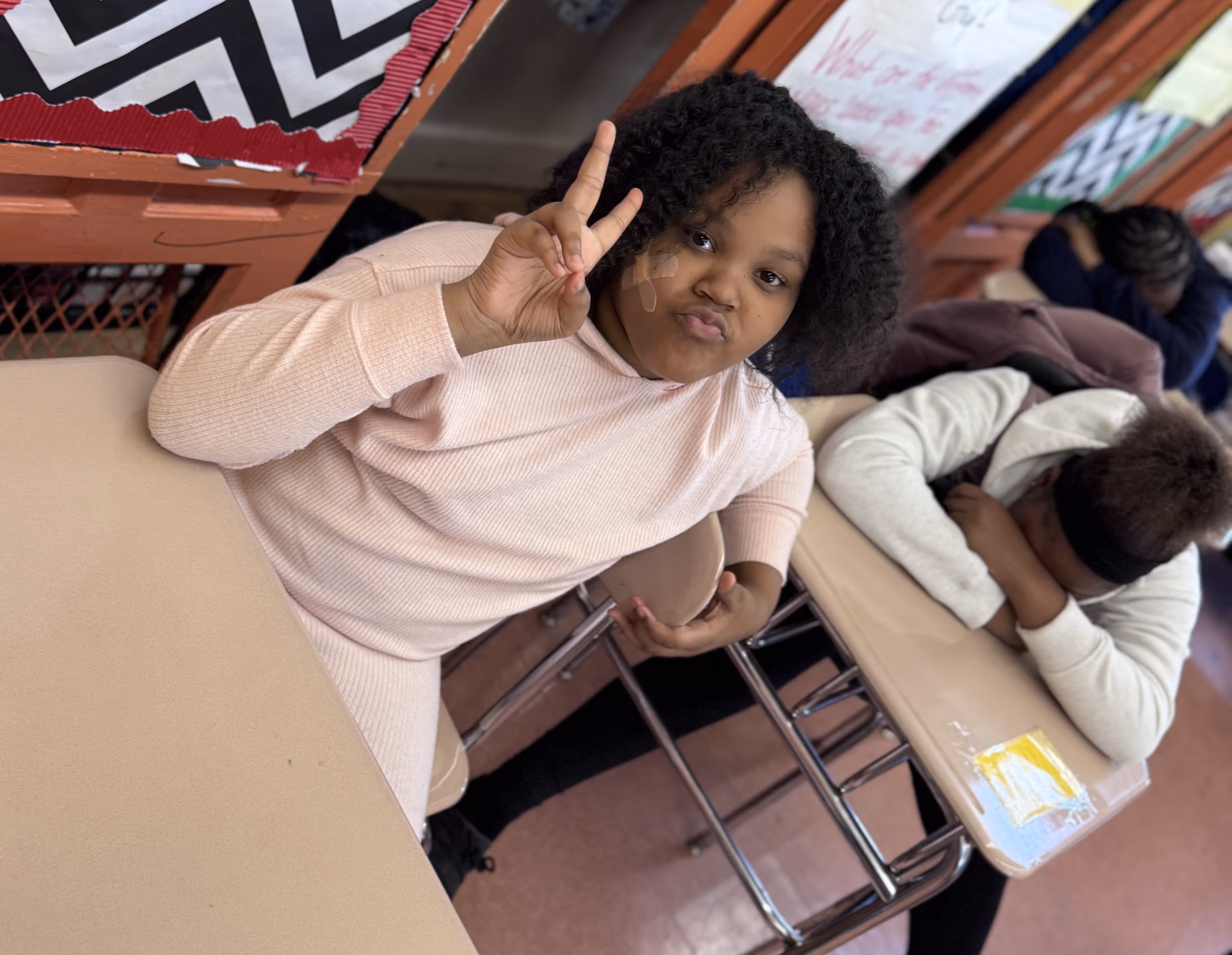 A young girl sitting at a classroom desk makes a peace sign and smiles playfully at the camera. She wears a light pink outfit. Behind her, two other students are resting their heads on their desks. The background features colorful posters and zigzag-patterned decorations.