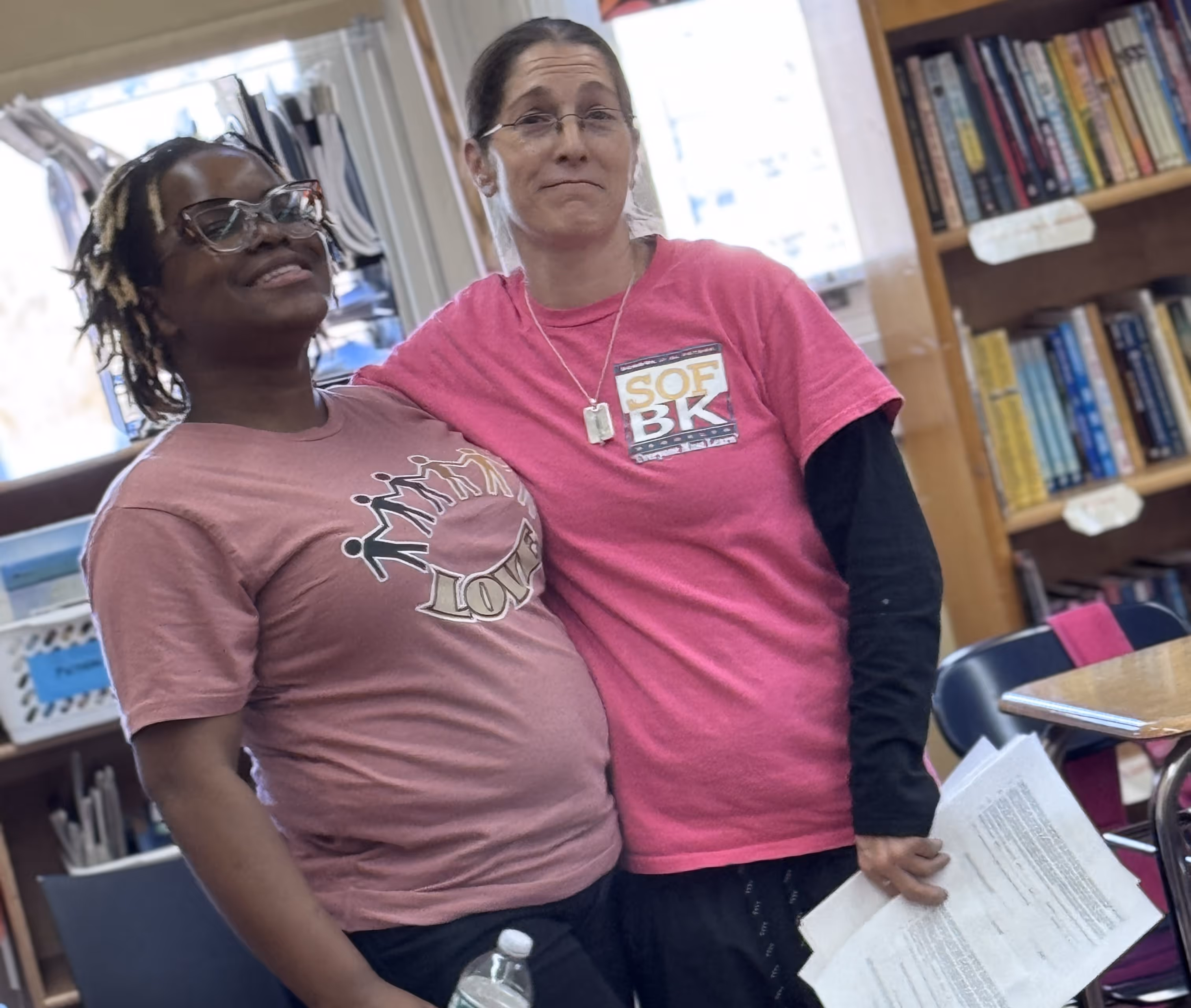 Two women stand side by side in a classroom, smiling and posing for the camera. One wears a shirt with the word “LOVE” and the other wears a bright pink shirt with “SOF BK” printed on it. Both appear friendly and supportive, with shelves of books visible in the background.

