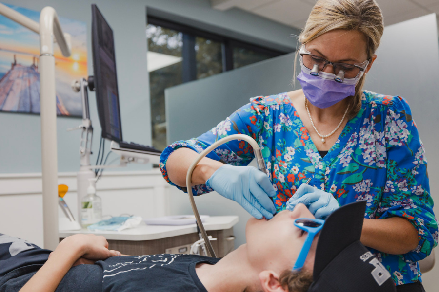 A patient wearing sunglasses and a baseball cap reclines in a dental chair while the orthodontist, dressed in a floral top and protective gear, carefully performs a procedure. A computer monitor and dental equipment are visible in the background.