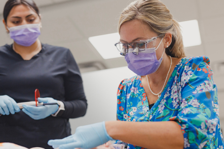 A female orthodontist wearing a floral blouse, face mask, and magnifying loupes focuses intently while working on a patient. A dental assistant stands nearby, holding an orthodontic tool, both wearing gloves and protective gear in a bright clinical setting.