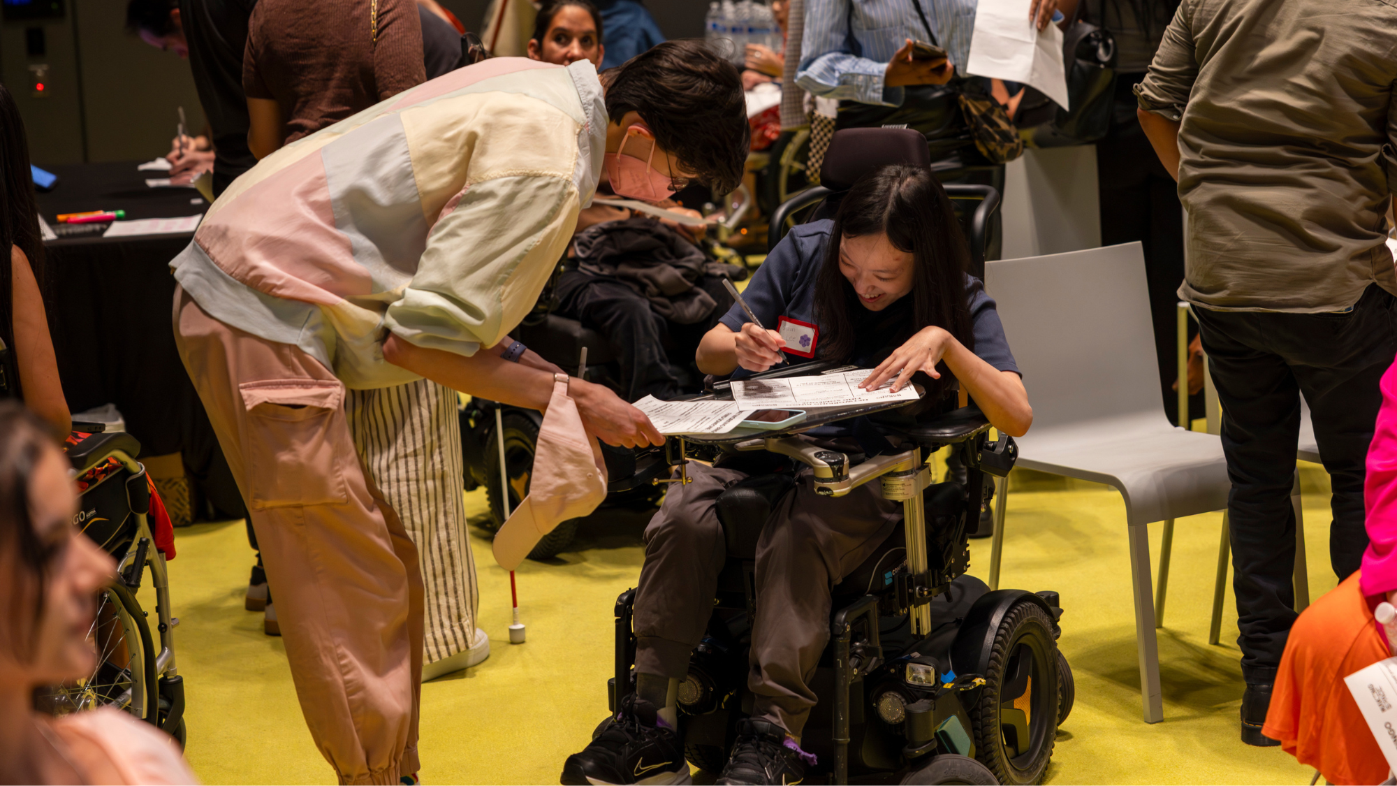 Image of Making Space community members bent over a worksheet at our Disability Pride Month event. One community member is standing and wearing a pink mask, multicolored windbreaker, and pink joggers. The other community member is using an electric wheelchair wearing a blue top and brown joggers. Photo was taken by Salvador Ceja Garcia. 