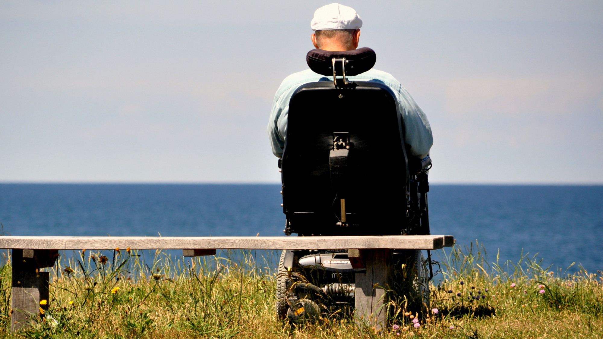 Picture of an electric wheelchair user on the grass looking out at a large body of water. 