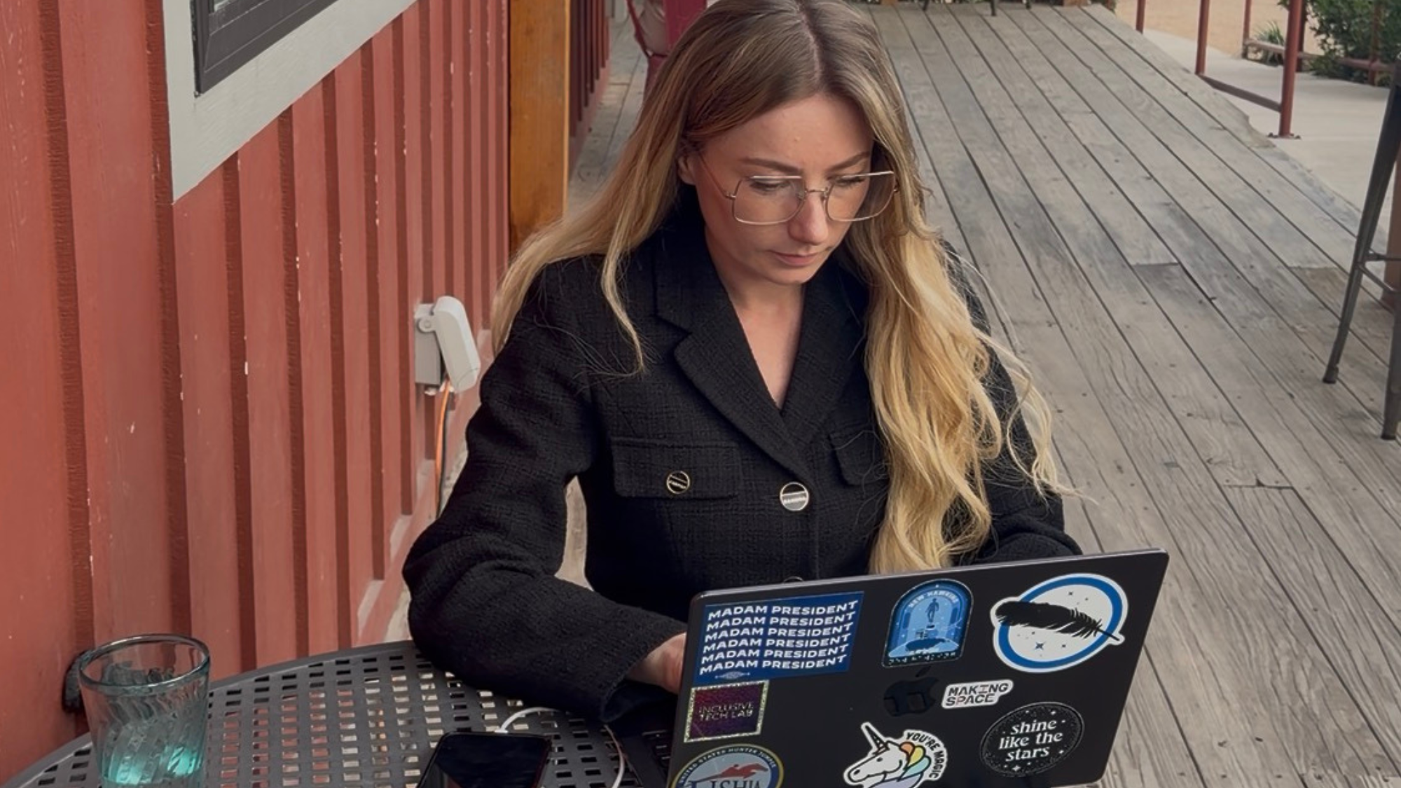 Photo of Keely working on her laptop at a coffee shop. Keely is a white woman with long blonde wavy hair. She is wearing large rectangular glasses and a black blazer.