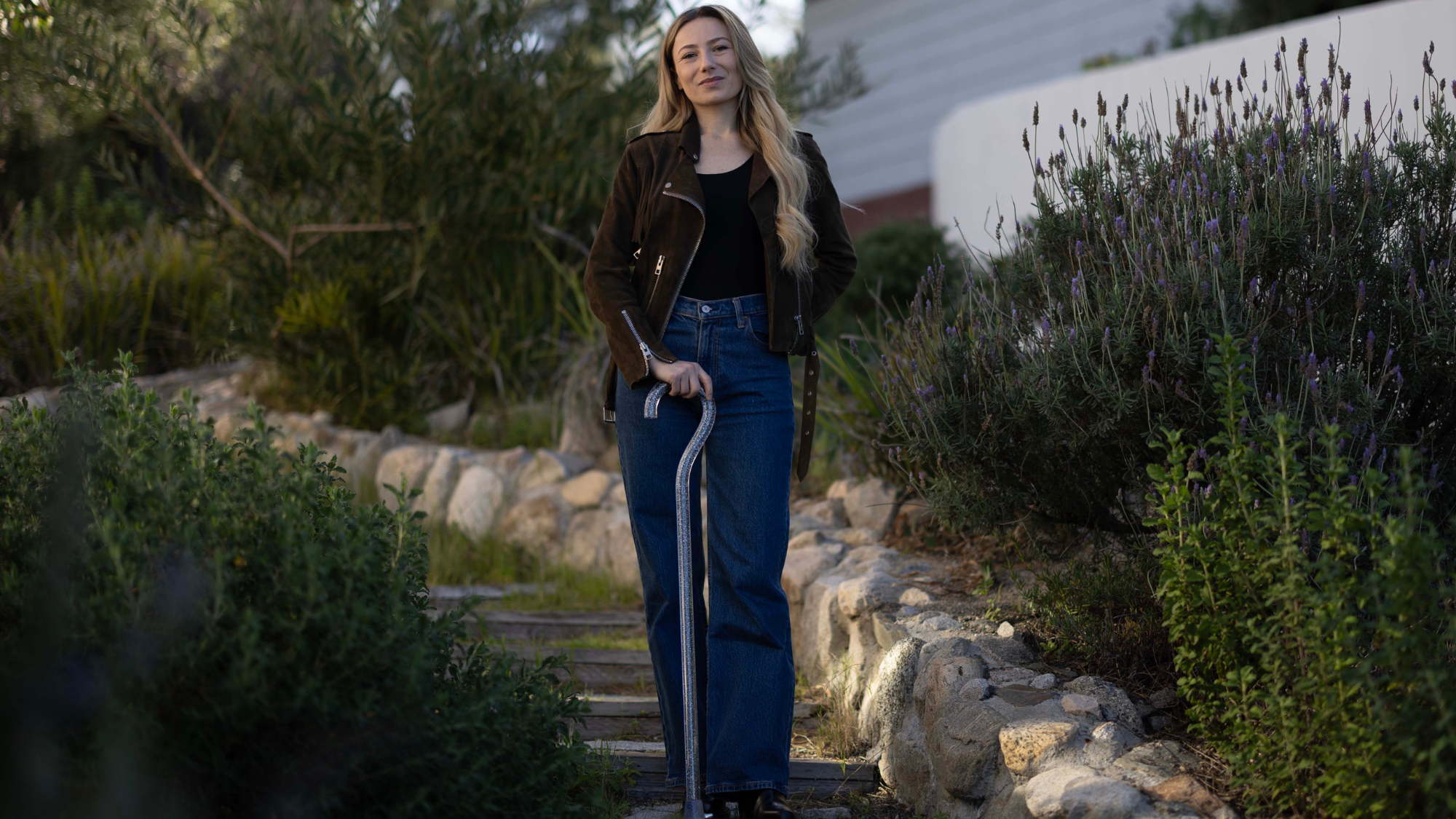 Photo of Keely standing on steps in a garden using her cane. She is wearing a black top, brown jacket, and blue jeans.