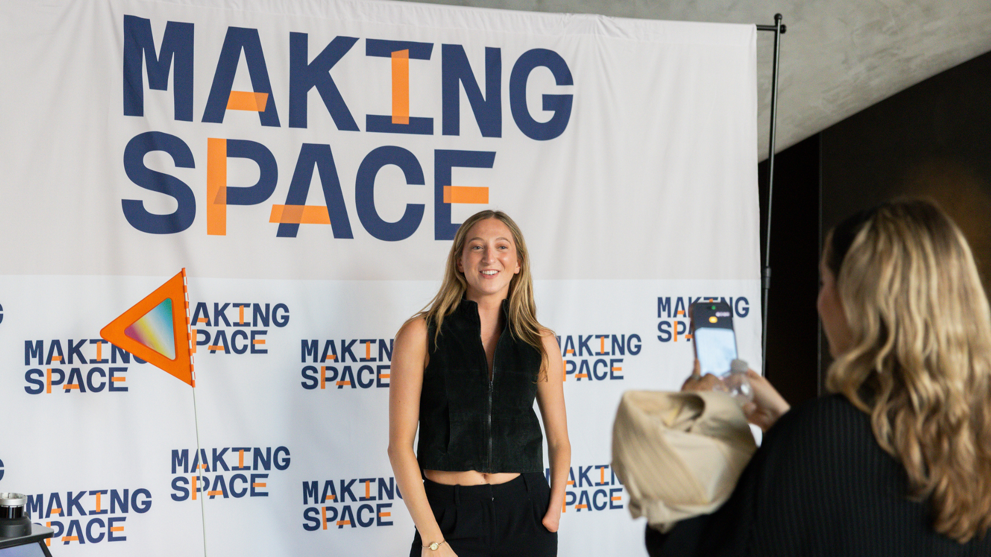 A woman with an arm limb difference is wearing a black sleeveless top and black pants and posing and smiling in front of a banner reading "Making Space" in navy and orange text. A second person stands to the right photographing them.