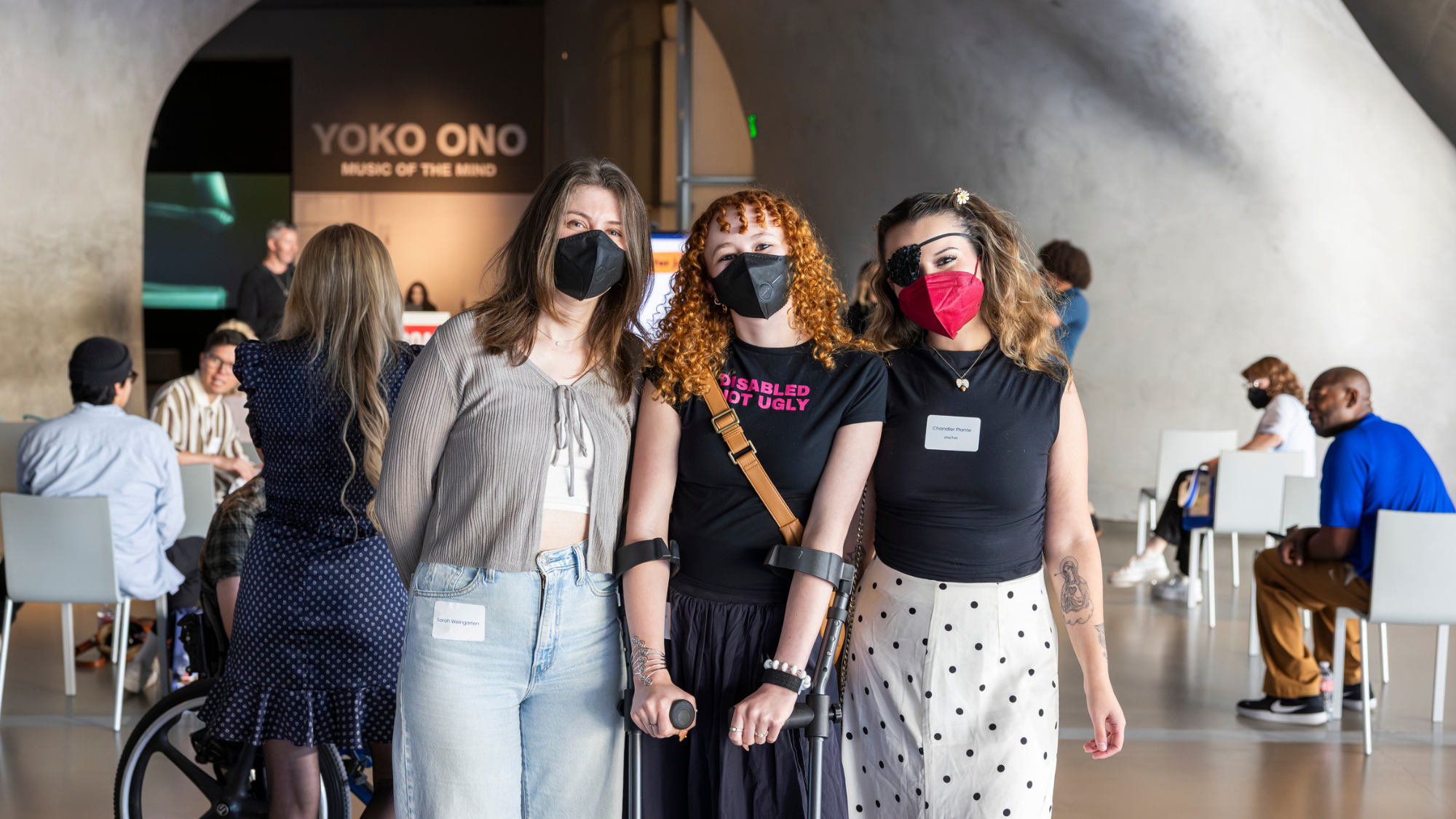 Three women wearing black face masks pose together and smile at the camera inside a museum. The center person is using forearm crutches and wearing a shirt that reads "Disabled Not Ugly." Behind the people posing is a sign for a Yoko Ono exhibition.