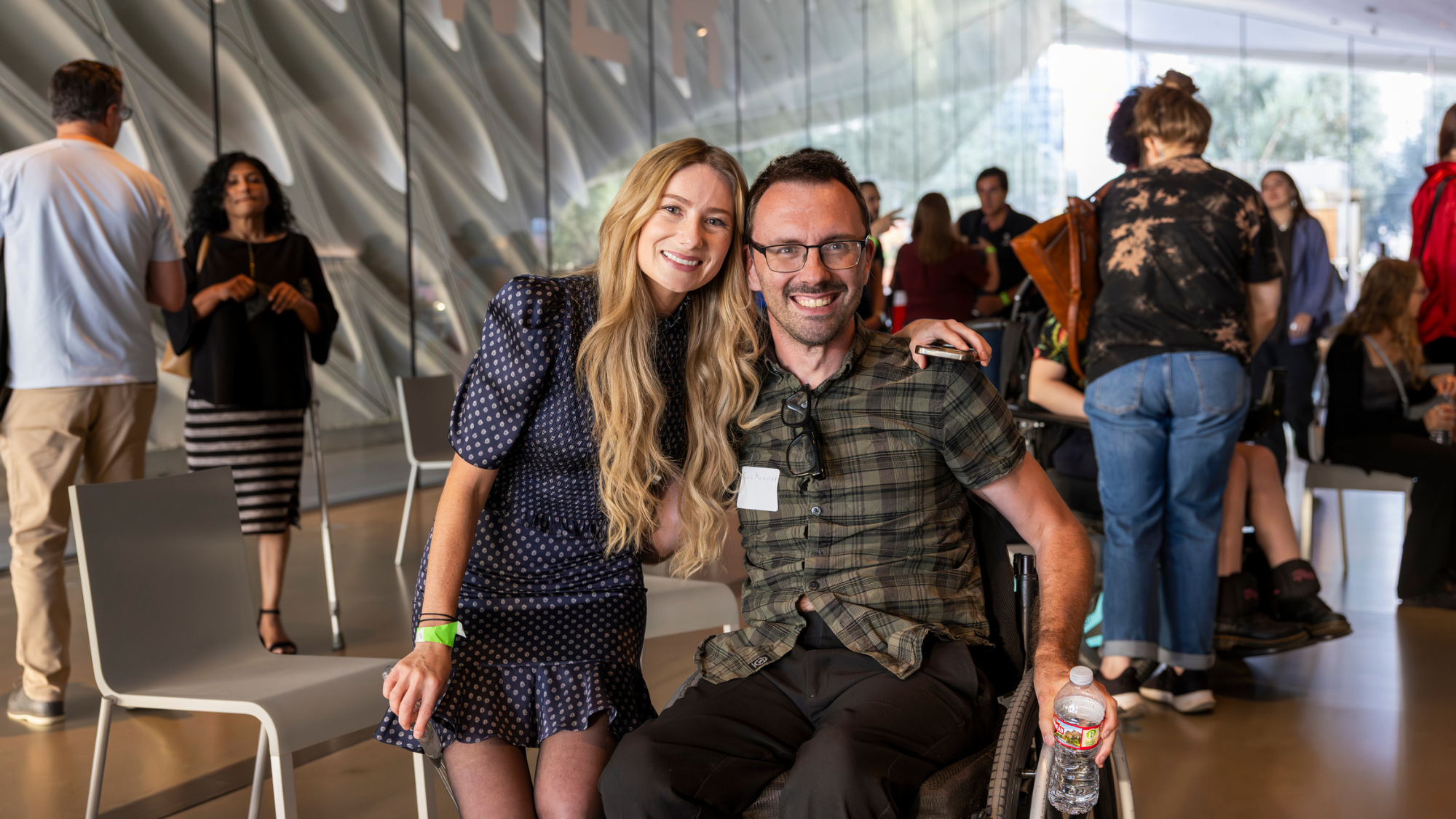 Keely, a woman with long blonde hair in a navy polka-dot dress leans beside a man in glasses who uses a wheelchair. In the background, a diverse crowd of attendees mingles in a museum setting.