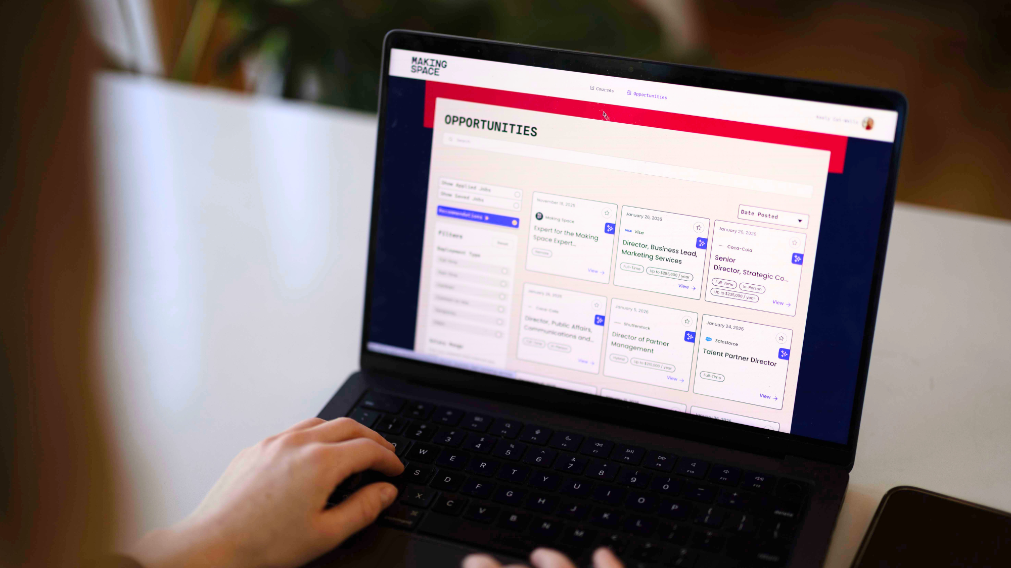Keely’s hands resting on a laptop keyboard while browsing the Making Space Opportunities page, which displays a grid of remote job listings from companies including Visa, Coca-Cola, Shutterstock, and Salesforce against a navy and bright pink branded interface.