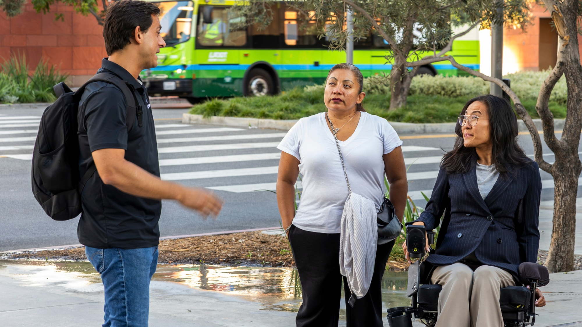 Three people congregate in an outdoor courtyard chatting. The courtyard is in front of a road with a bus passing through. On the left is a man standing with tan skin, brown short hair, a black polo shirt, blue jeans, and a black backpack. In the middle is a woman standing with tan skin, brown and grey hair pulled back in a low ponytail, she is wearing a white v neck top, black pants, a black crossbody purse and red lipstick. On the right is an Asian American woman using a power wheelchair. She has medium length wavy black hair and is wearing glasses, a double breasted pin striped blazer, and khaki pants.