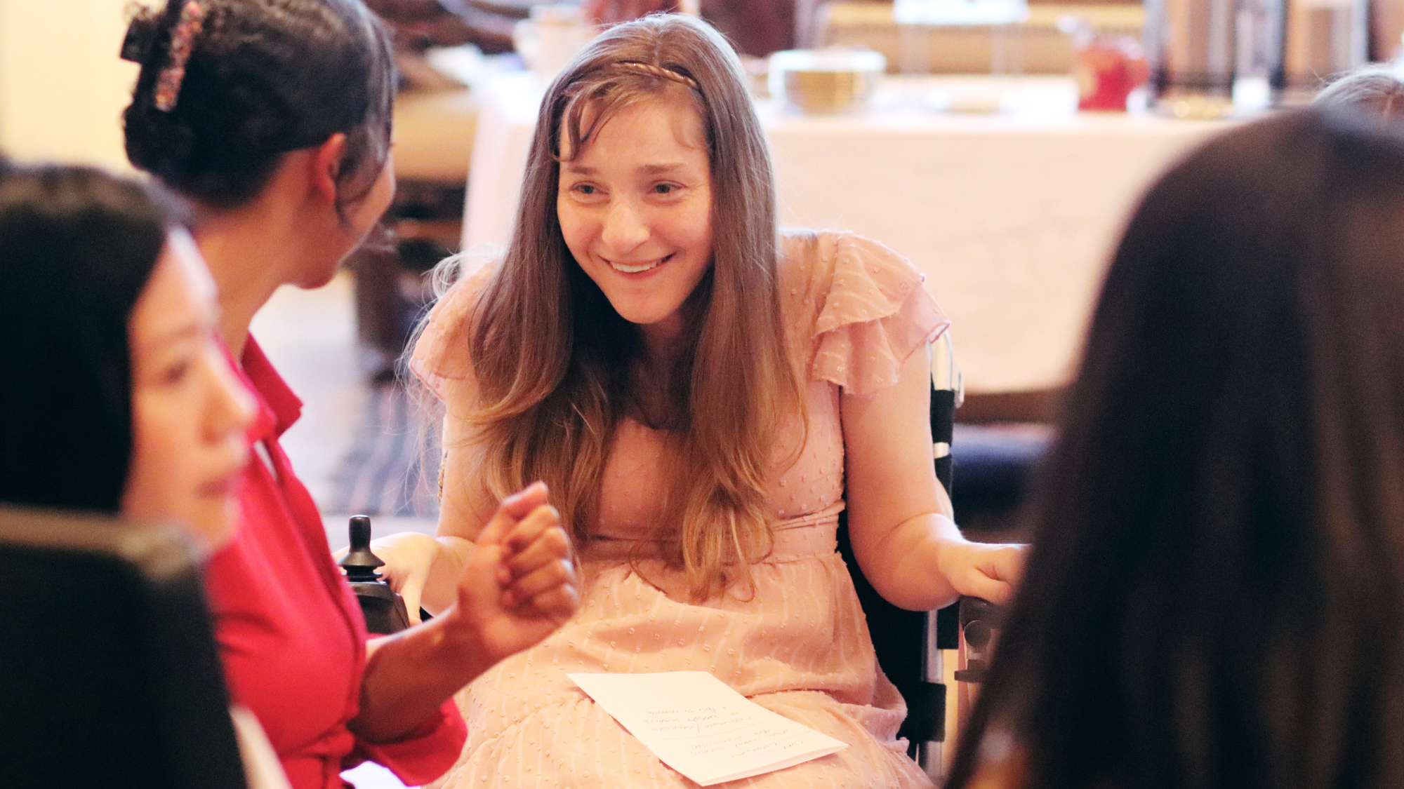 A white woman with long brown hair in a pink ruffled dress uses a power wheelchair, and smiles warmly while holding a piece of paper and engaging in conversation with 2 other people.