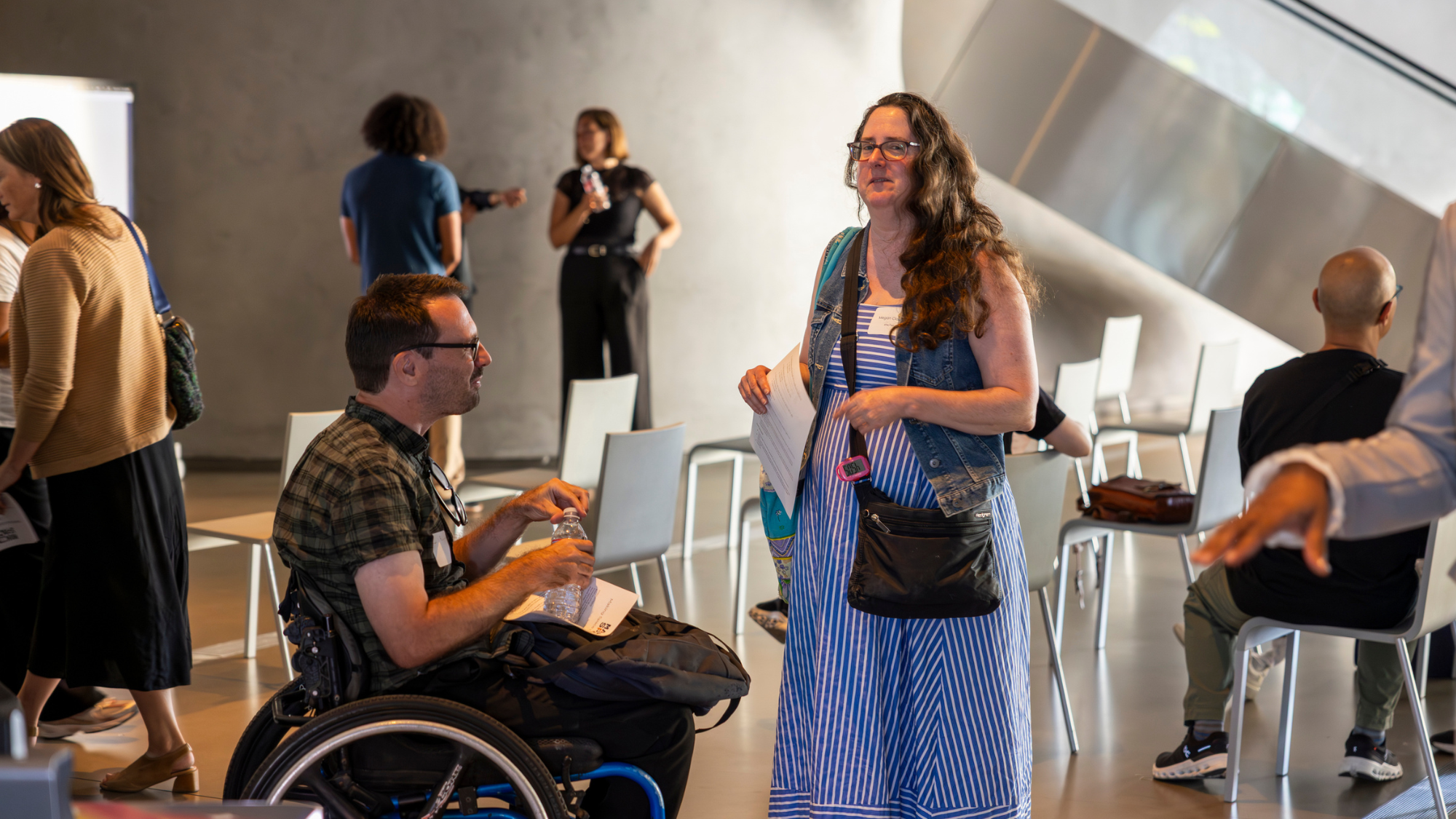 A person who appears to be a white man in a plaid shirt and glasses, seated in a wheelchair and holding a water bottle, chats with a person who appears to be a white woman in a blue striped dress and denim vest who stands holding papers. Around them, other attendees mingle and sit among white chairs in a bright, modern event space.