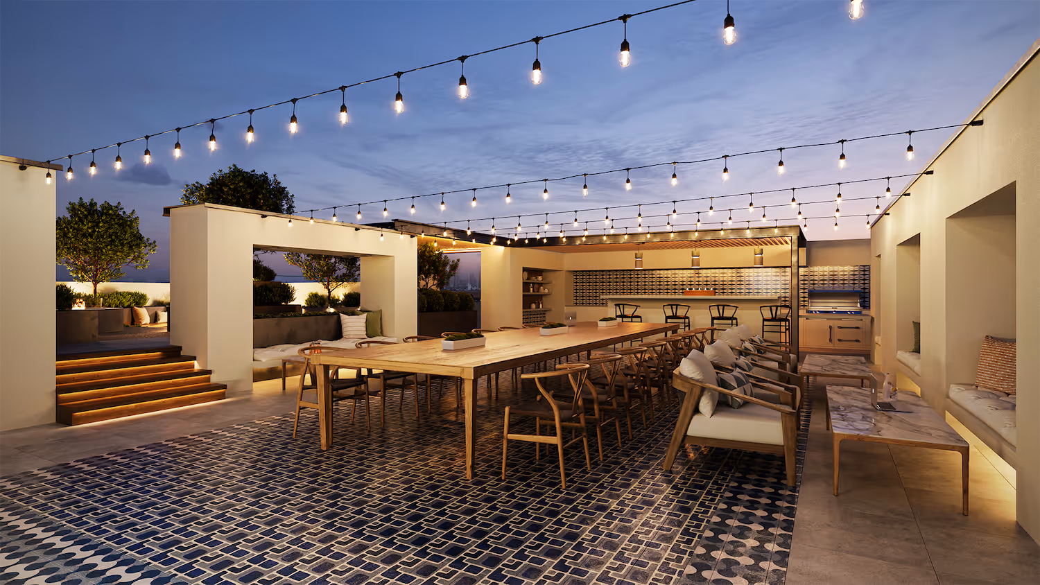 Rooftop dining area with a long table, chairs, and an outdoor kitchen lit by string lights at night, a stylish amenity at Tessera in Los Angeles, CA.