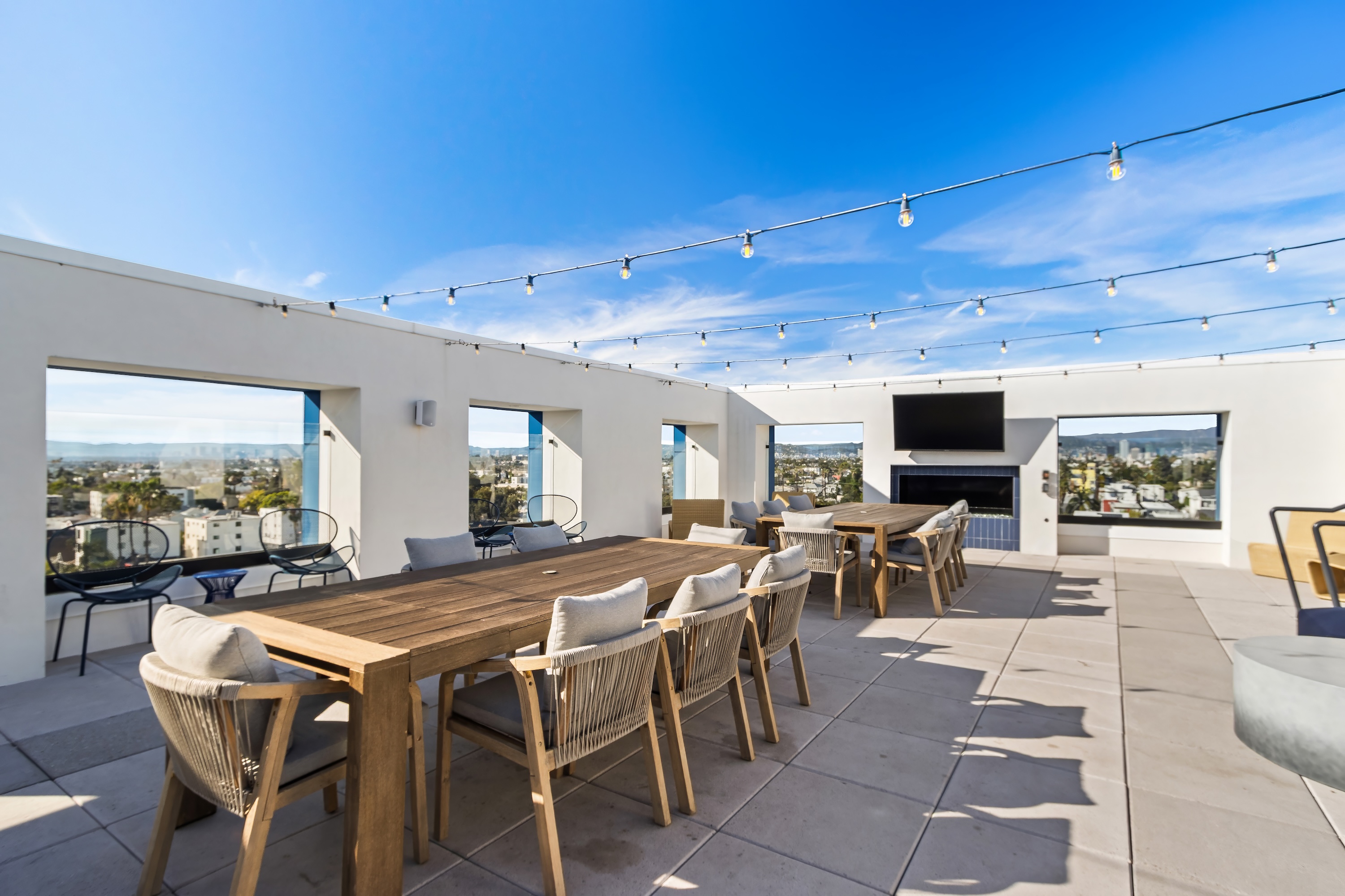 Rooftop dining area with a long table, chairs, and an outdoor kitchen lit by string lights at night, a stylish amenity at Tessera in Los Angeles, CA.