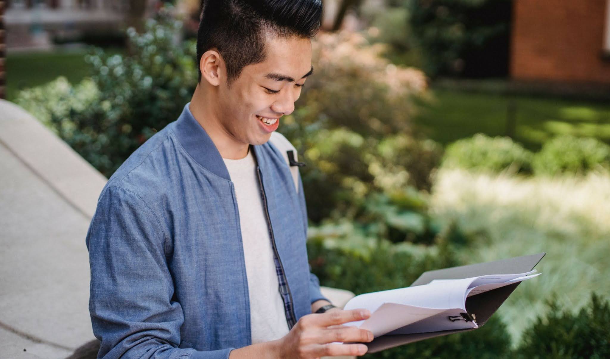 Person reading papers from a binder on a stone bench beside leafy garden shrubs