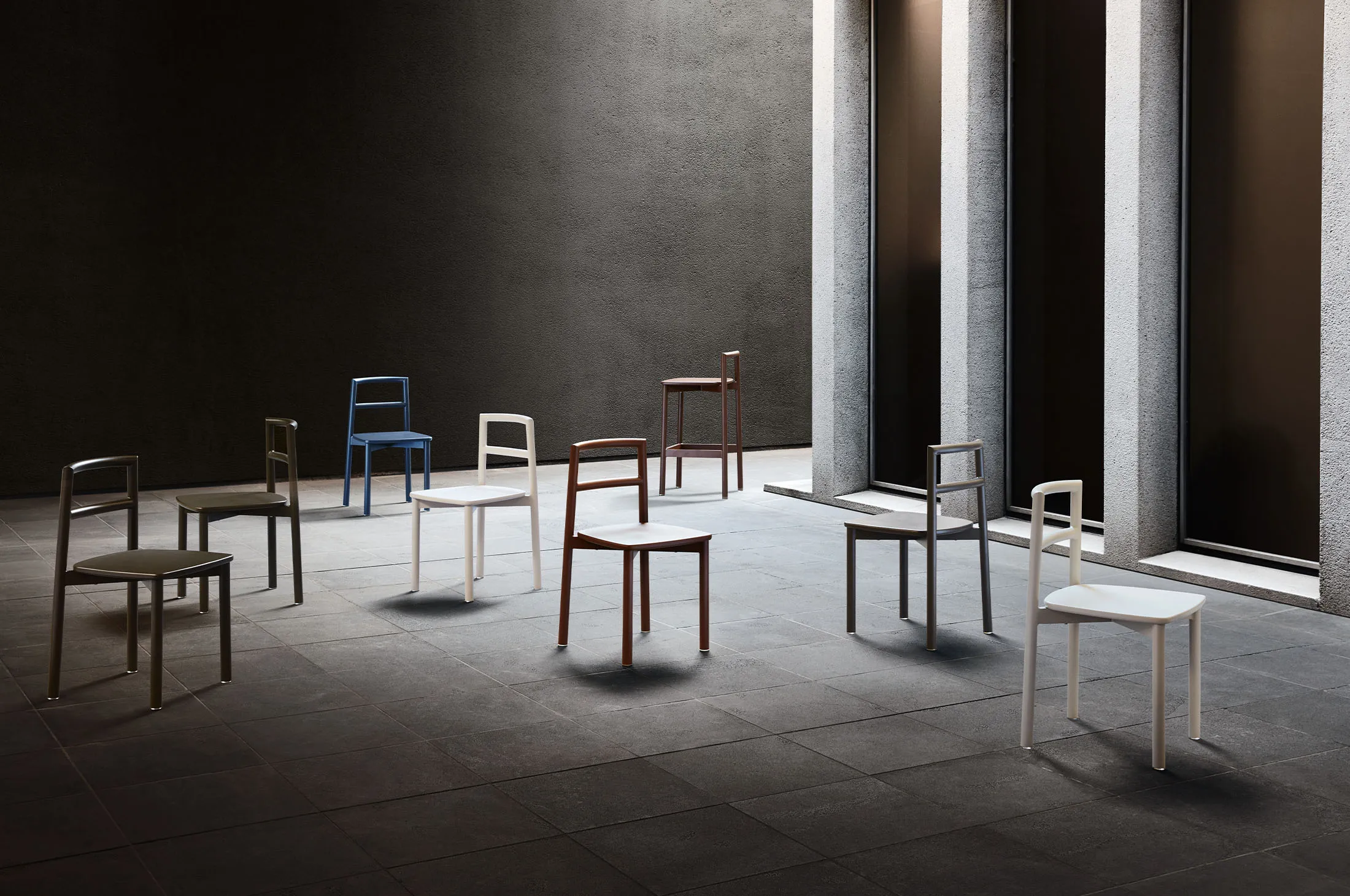 Modern minimalist chairs and stools in blue, brown, and white on gray tiled floor under concrete pillars.