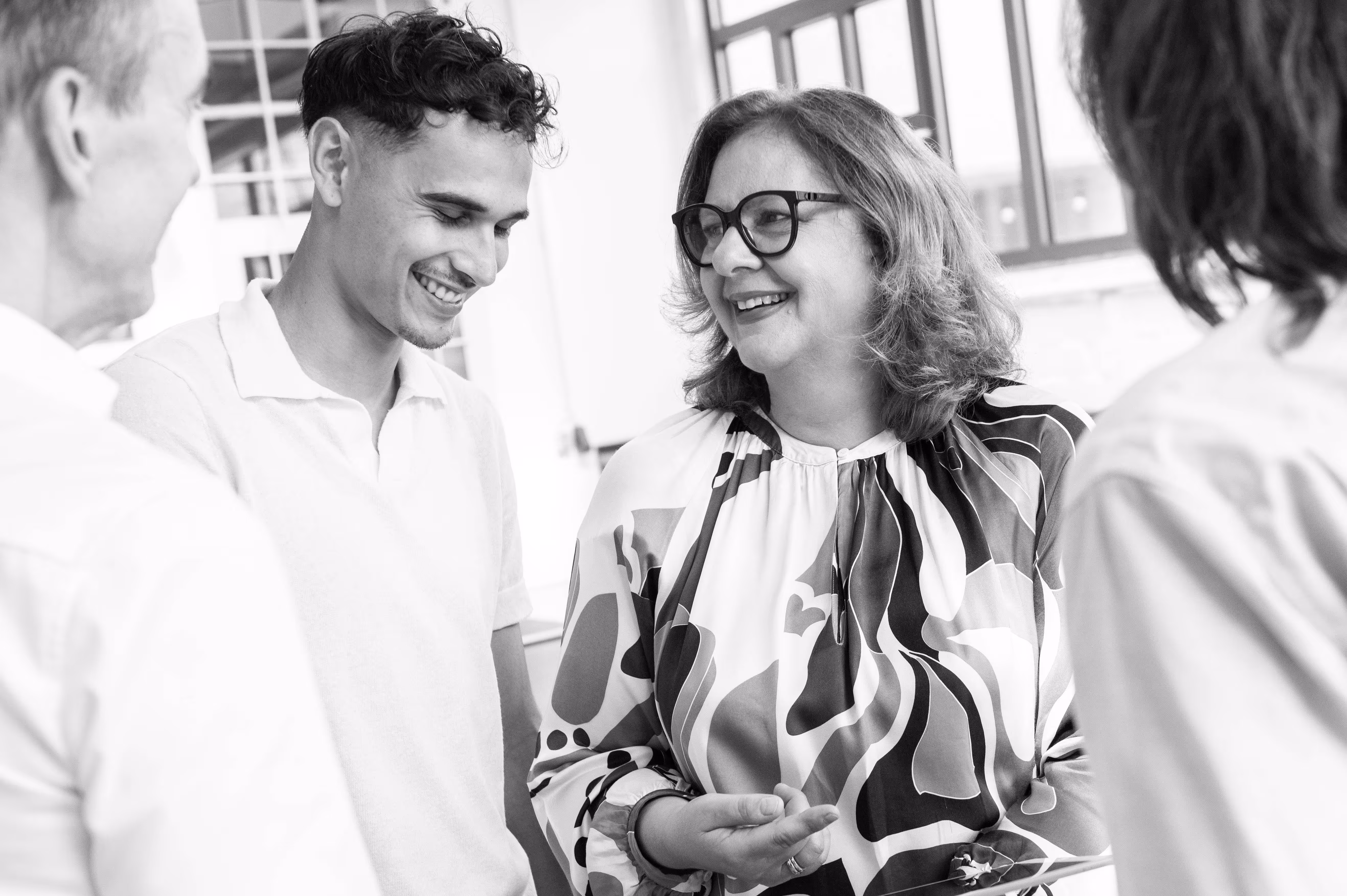 A woman with glasses and patterned blouse smiling and talking with three people in a bright room.