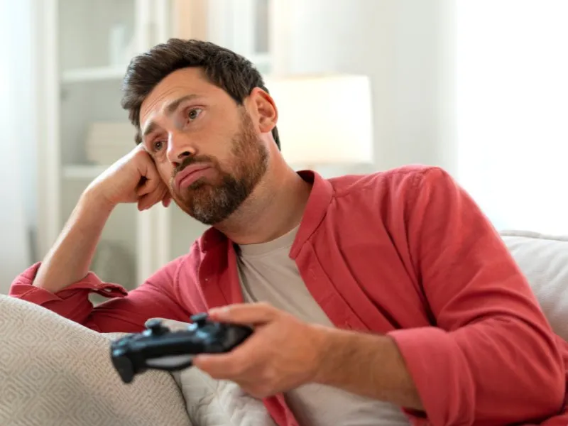 A middle-aged man wearing a red-orange coat grips a PlayStation controller, his expression marked by frustration and disengagement, suggesting he's not enjoying the experience.