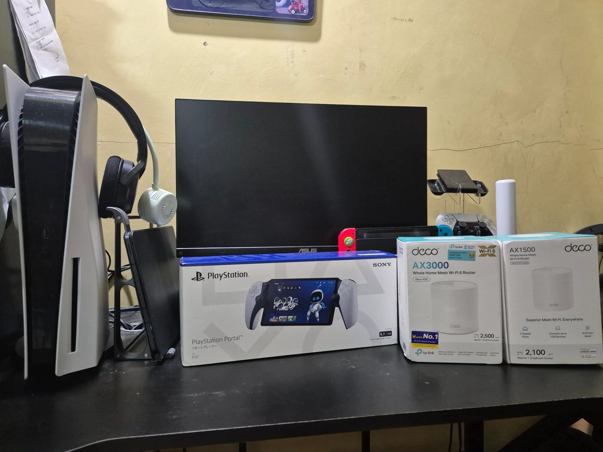 A table that shows the following: PS5 console at the left, a box of PS Portal in the middle, Deco mesh wifi routers on the right, and a monitor and Nintendo Switch at the background