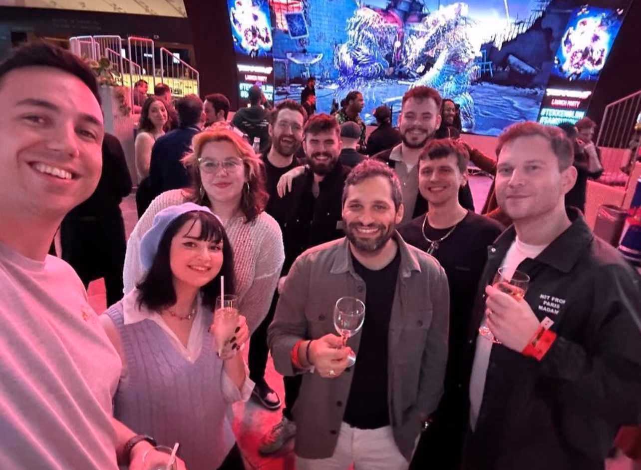 Group of nine friends smiling and holding drinks at a lively indoor event with colorful lighting.