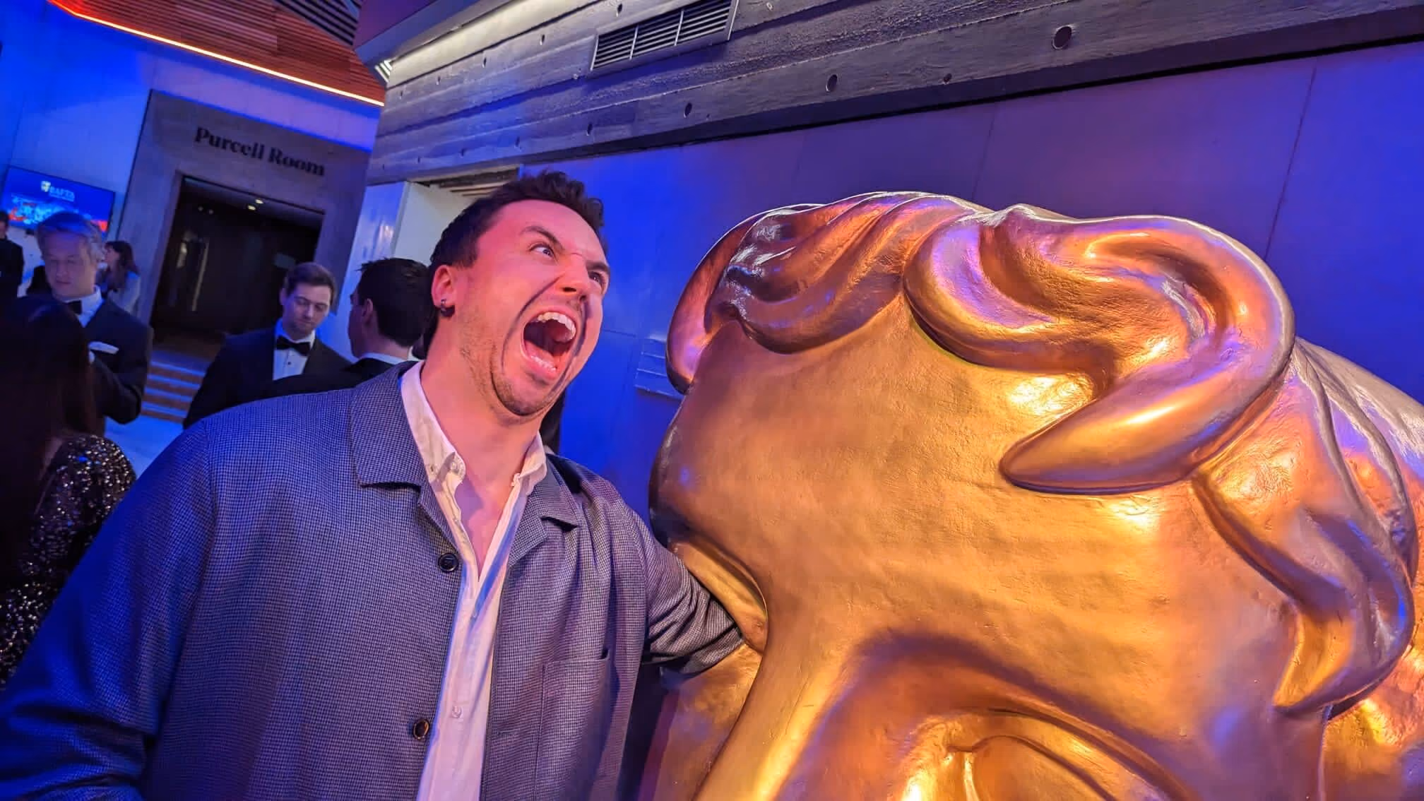 Man pretending to bite a large golden statue head at a formal event with blue lighting.