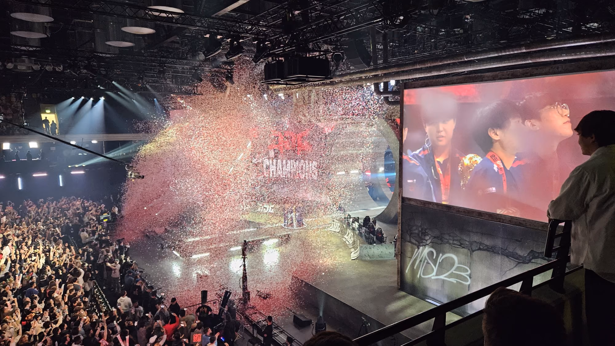 Crowded arena celebrating champions with confetti and large screen showing team with trophy.