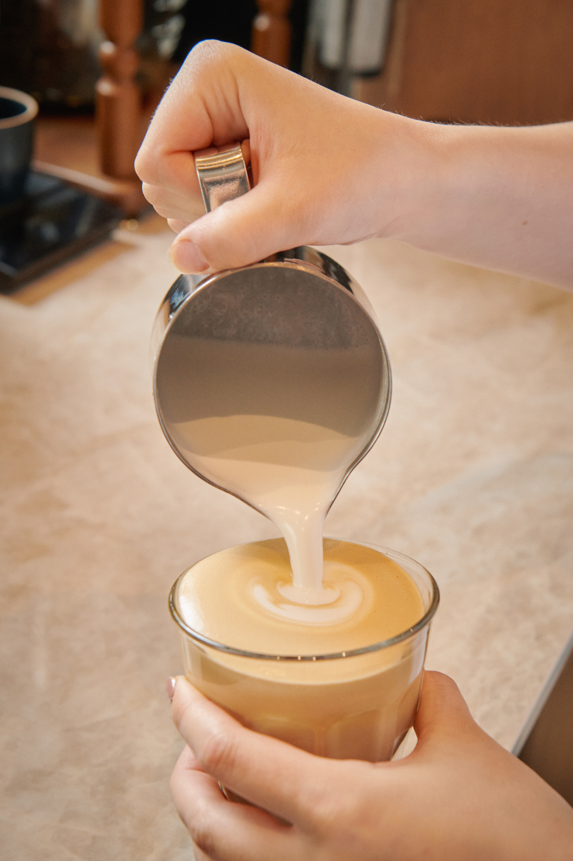 Person pouring steamed milk from a metal pitcher into a glass of coffee to create latte art.