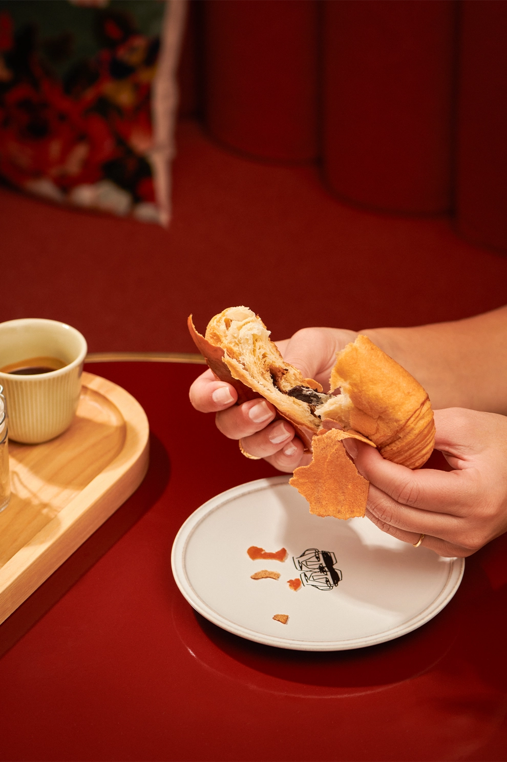 Hands breaking a chocolate-filled croissant over a white plate with crumbs on a red table next to a wooden tray holding a cup of coffee.