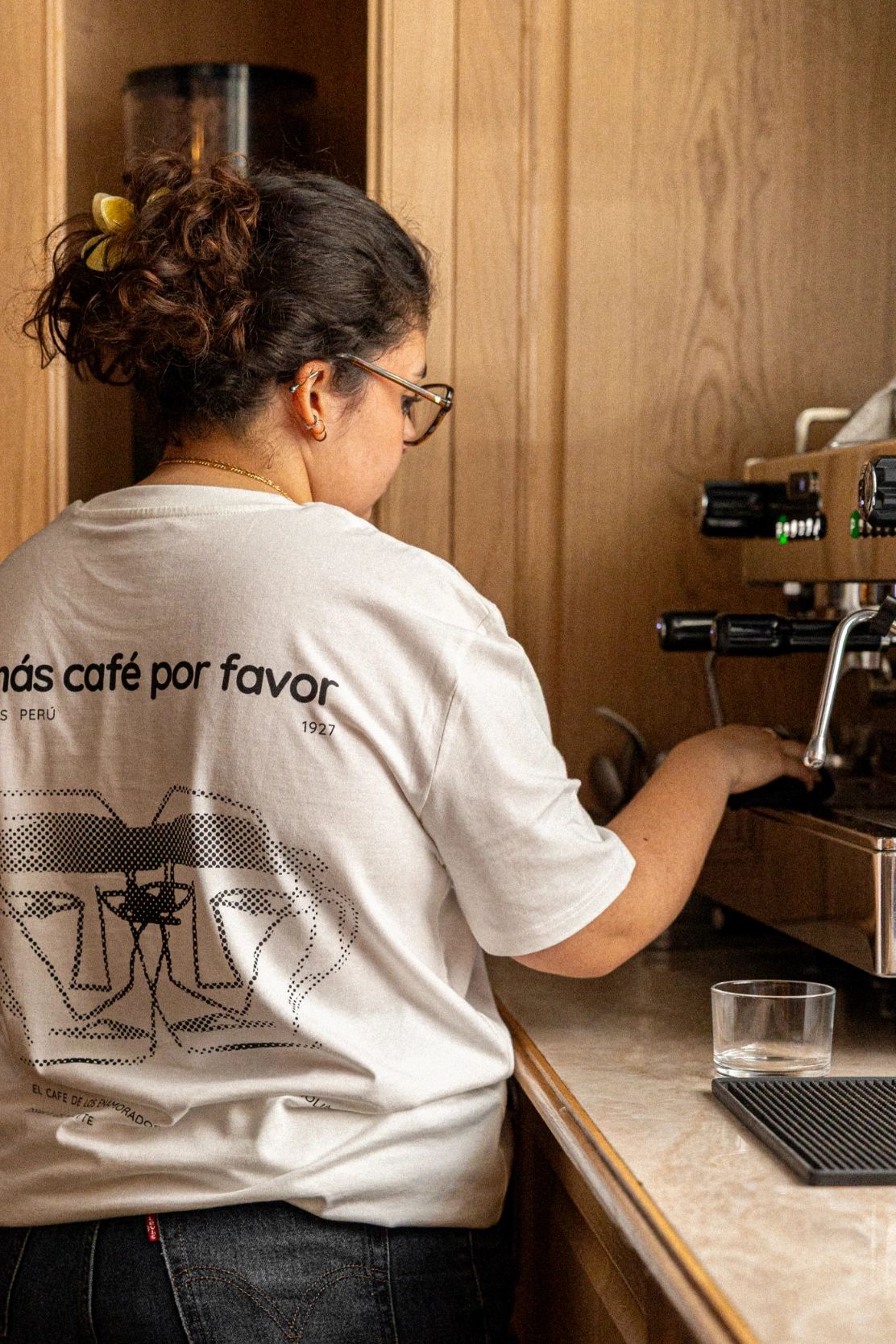Woman with curly hair tied with a yellow clip, wearing glasses and a white shirt, preparing coffee on an espresso machine in a wooden interior.