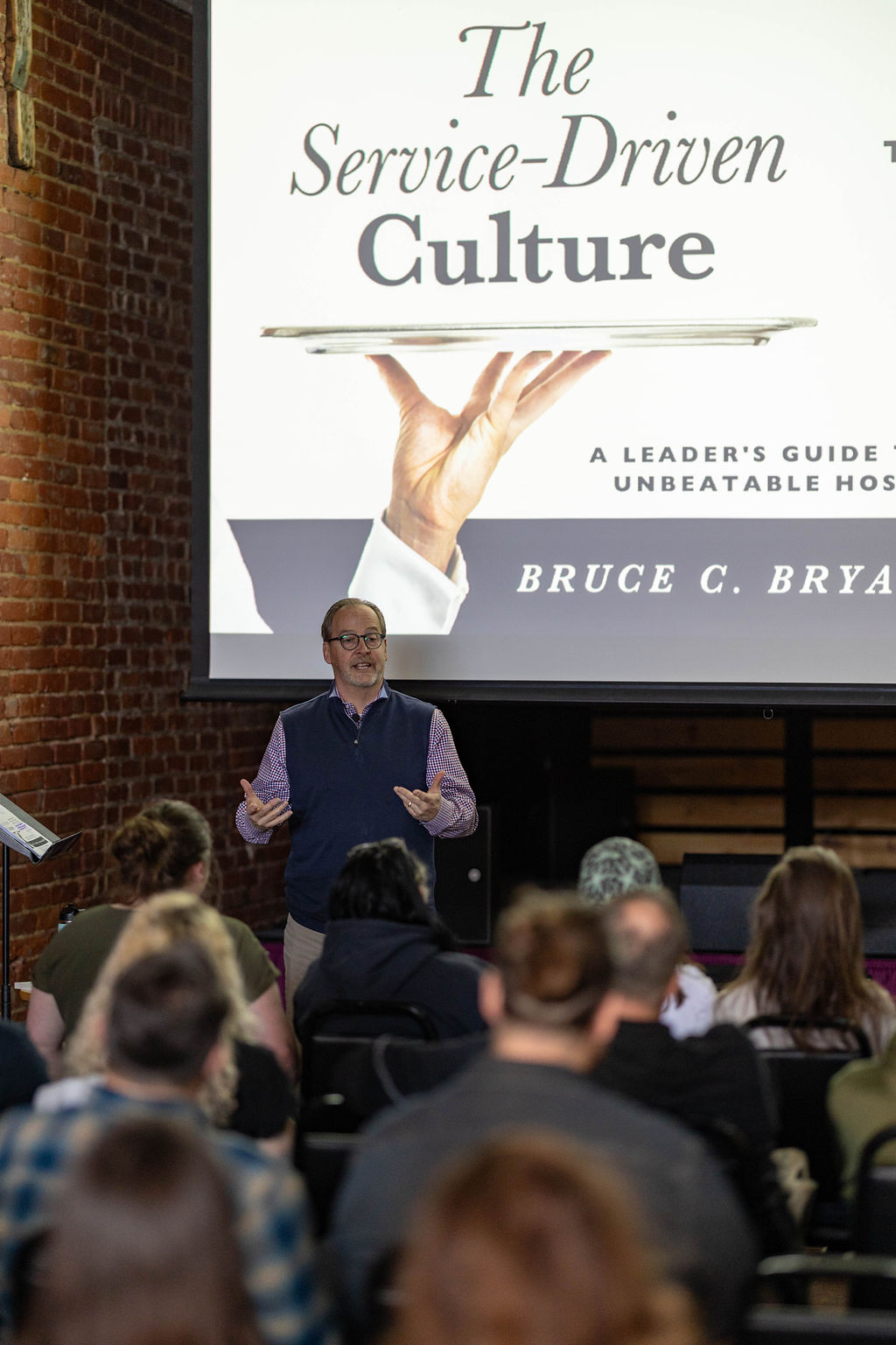 Person addressing a seated audience in an indoor space with exposed brick walls, standing in front of a large presentation screen that reads “The Service‑Driven Culture.”