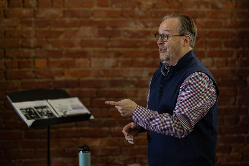 Person presenting indoors, gesturing with one hand near a music stand holding printed materials, set against an exposed brick wall.