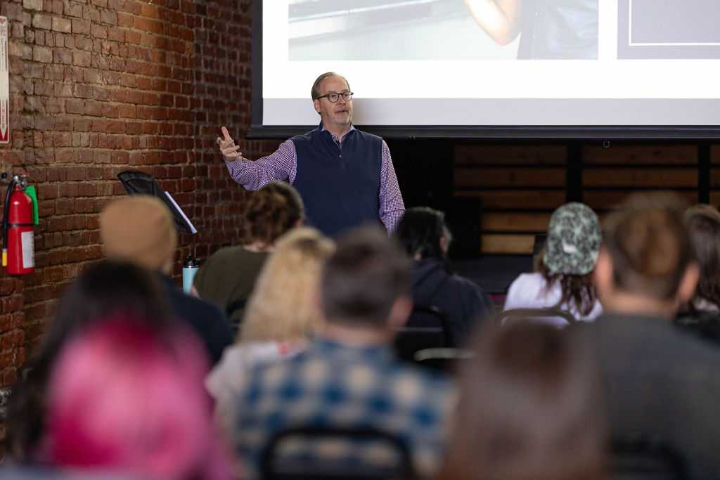Person speaking to a seated audience, gesturing with one hand in front of a presentation screen.