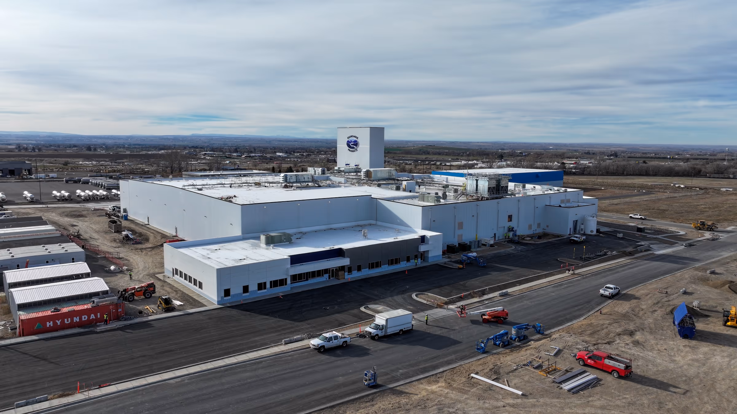 Aerial view of a large industrial building complex with white roofs, surrounded by trucks and construction equipment on paved and unpaved roads.